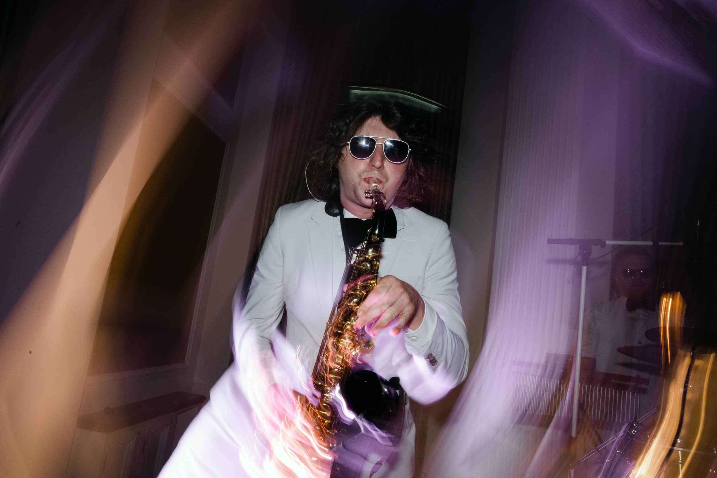 Person with curly hair wearing sunglasses and a white suit playing a saxophone, with colorful light streaks and blurred background.