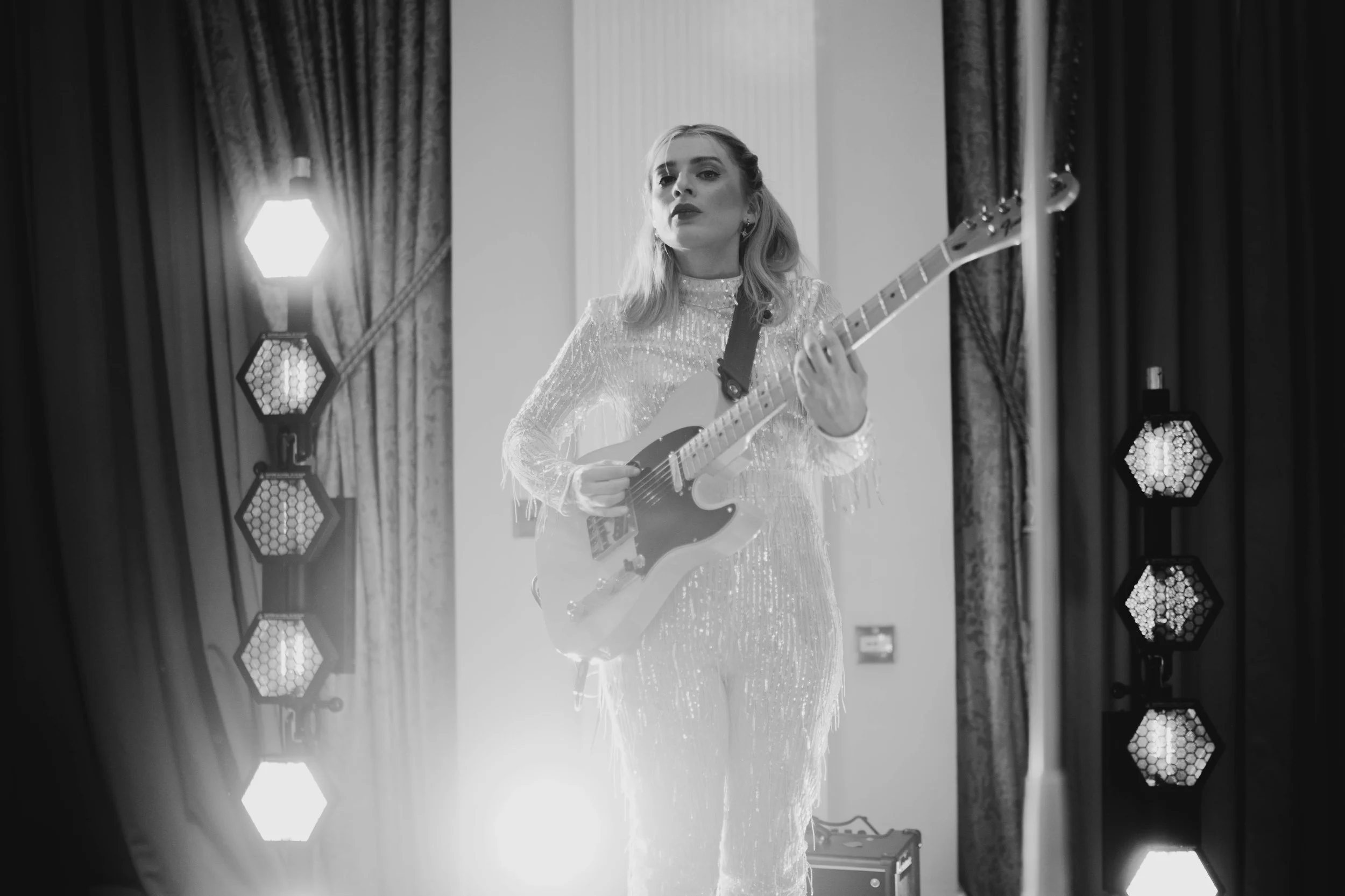 Black-and-white photo of a woman playing an electric guitar on a stage with curtains, stage lights, and equipment.