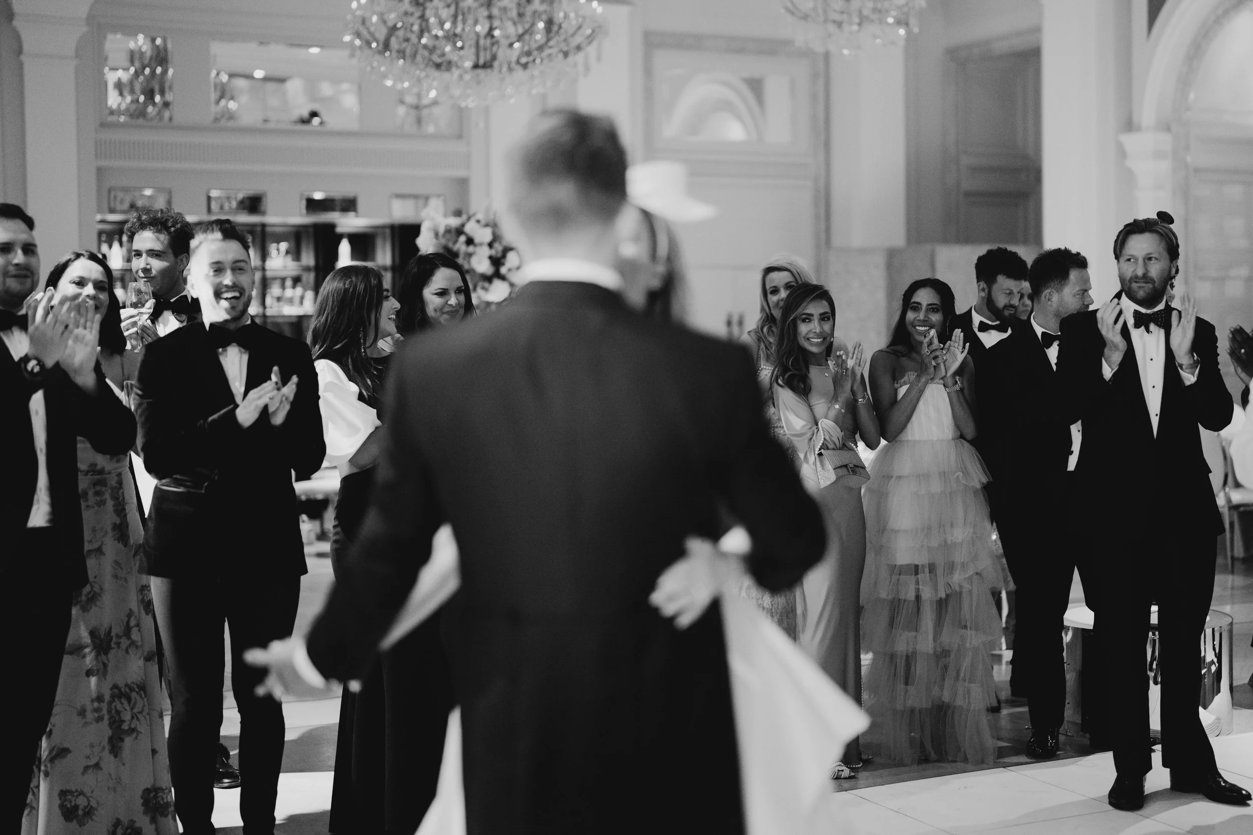People in formal attire at a wedding reception, clapping and smiling in a decorated elegant ballroom.