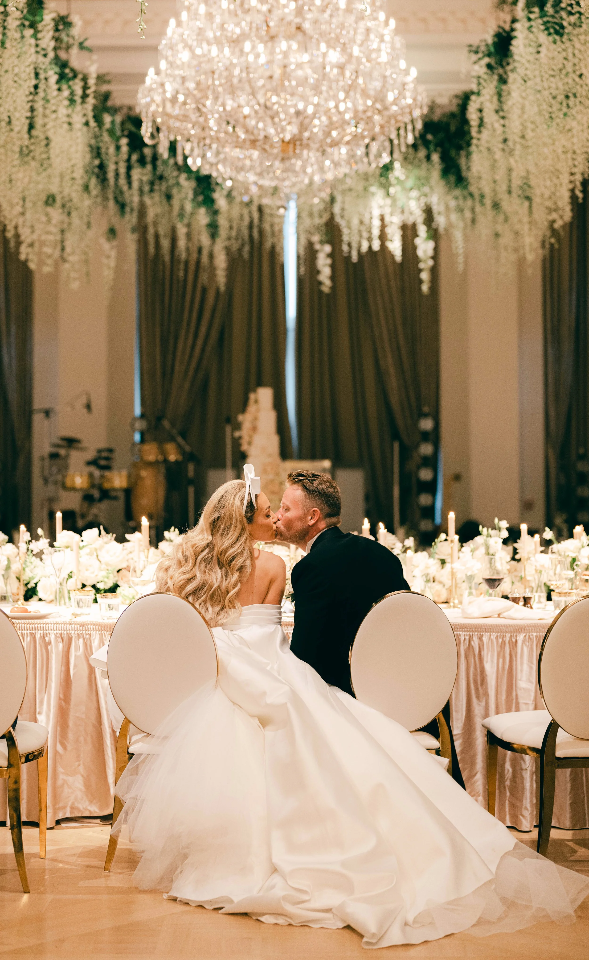 Bride and groom kissing at a lavish wedding reception, sitting on white chairs, under a large chandelier with floral decorations.