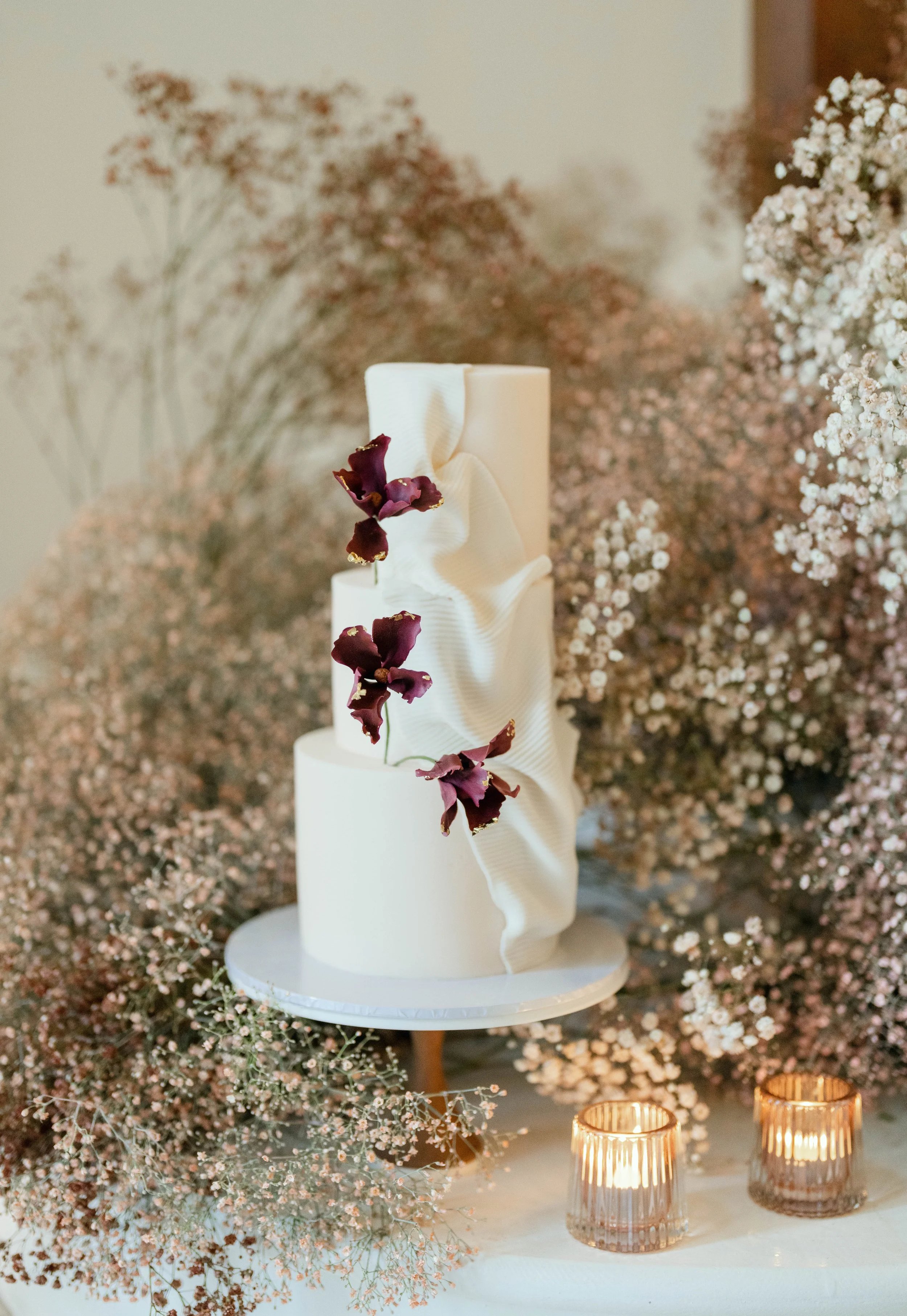 Elegant wedding cake with white fondant, adorned with purple flowers and draped white fabric, surrounded by pink baby’s breath flowers and two small lit candles.
