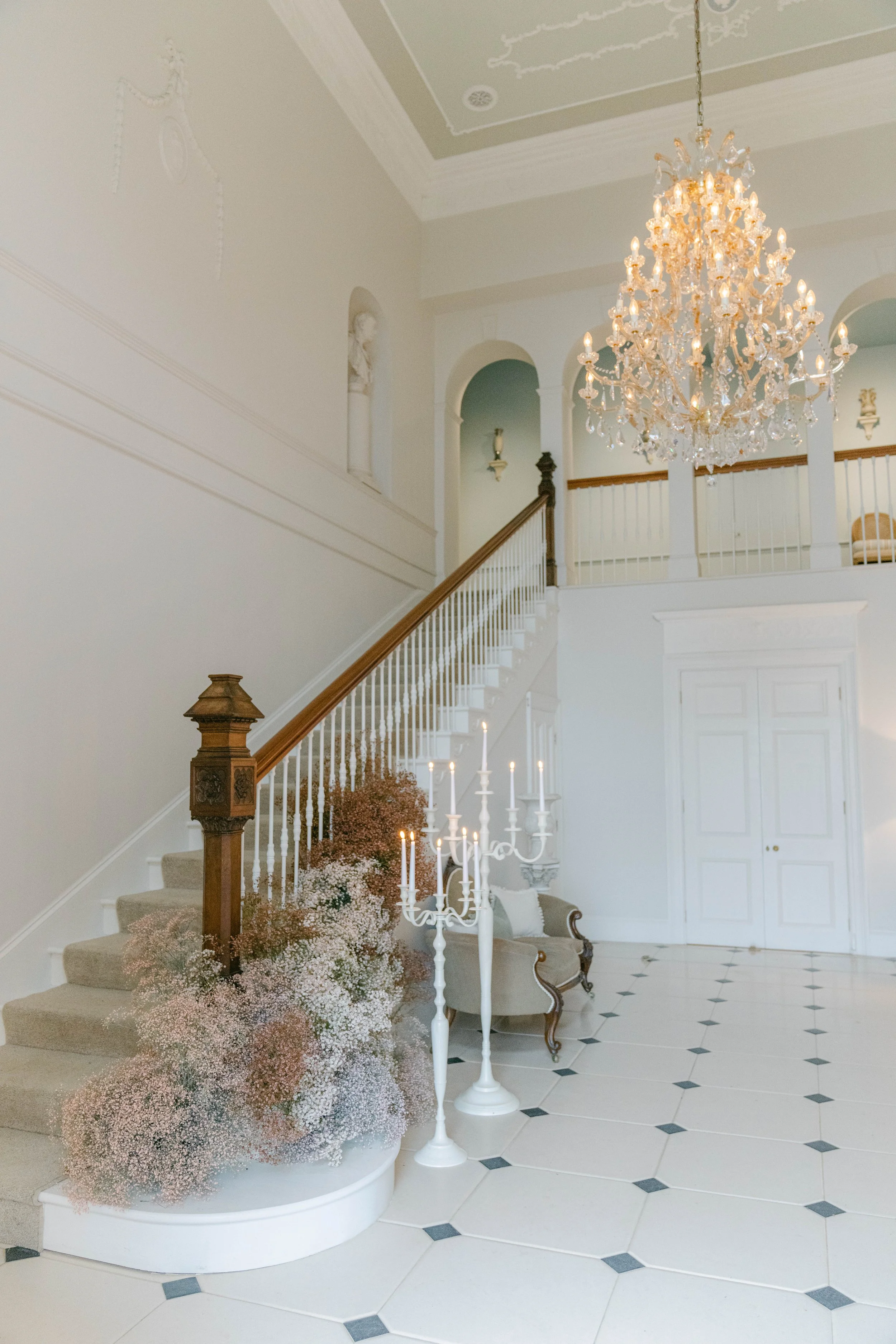 Elegant foyer with staircase, large chandelier, White candelabra, beige seating, floral arrangement, white and black tiled floor, decorative wall molding.