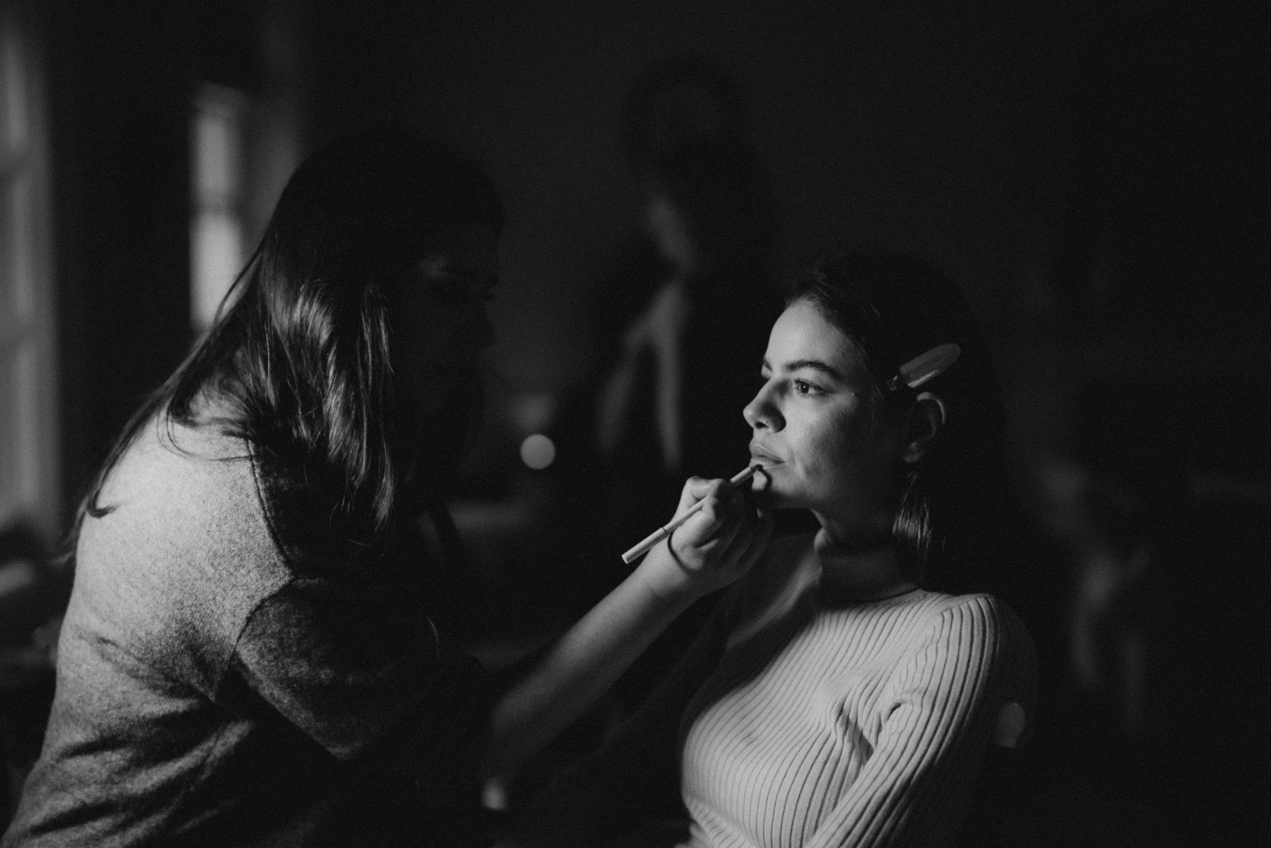 Black and white image of a makeup artist applying lipstick to a woman sitting in a chair, with soft lighting creating a shadowed ambiance.