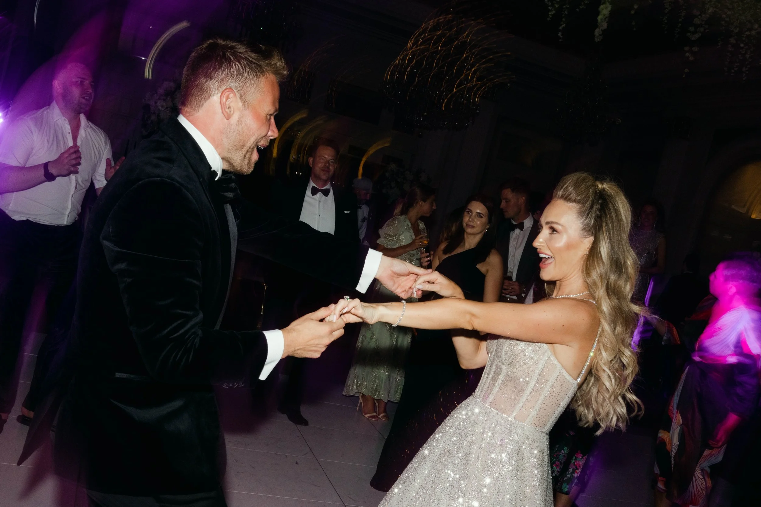A bride and groom dancing and holding hands at their wedding reception, surrounded by guests in formal attire.