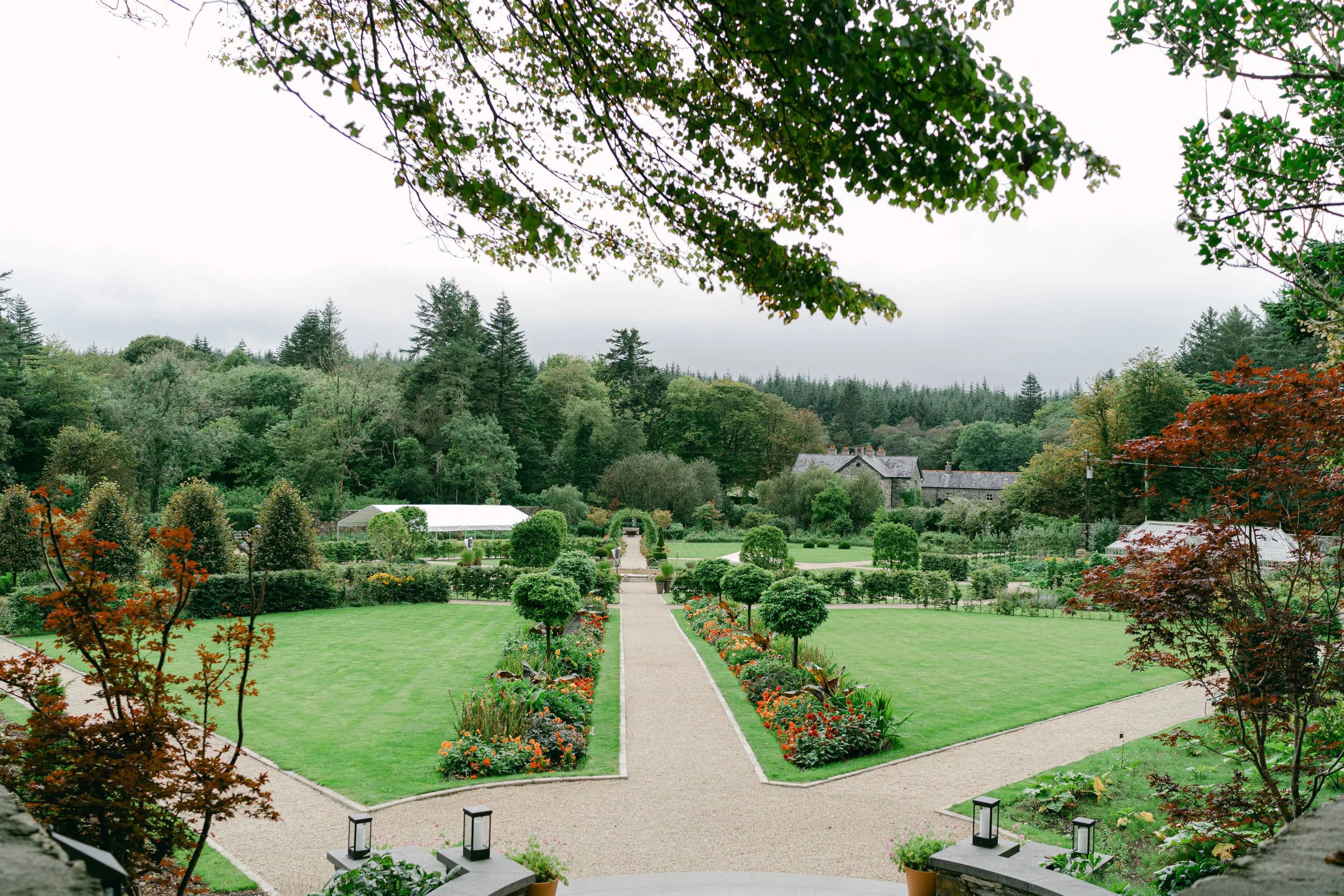 Formal garden with manicured lawns, flower beds, and hedges, set against a backdrop of dense woodland and a large house. Pathways lead through the garden, surrounded by vibrant greenery and colorful flowers.