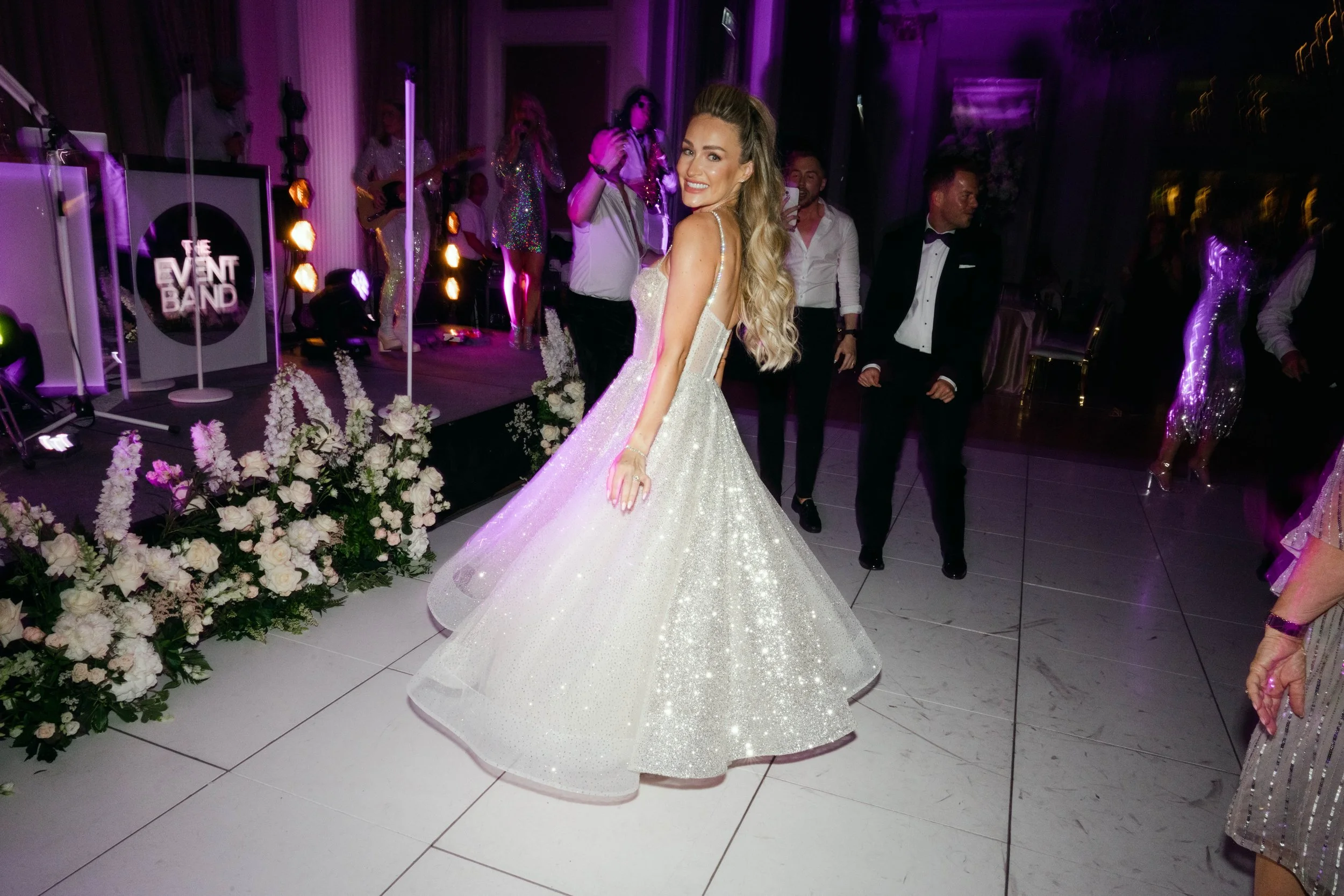 A woman in a sparkling white wedding gown dancing at a celebration, with a band playing on stage and guests in formal attire in the background.