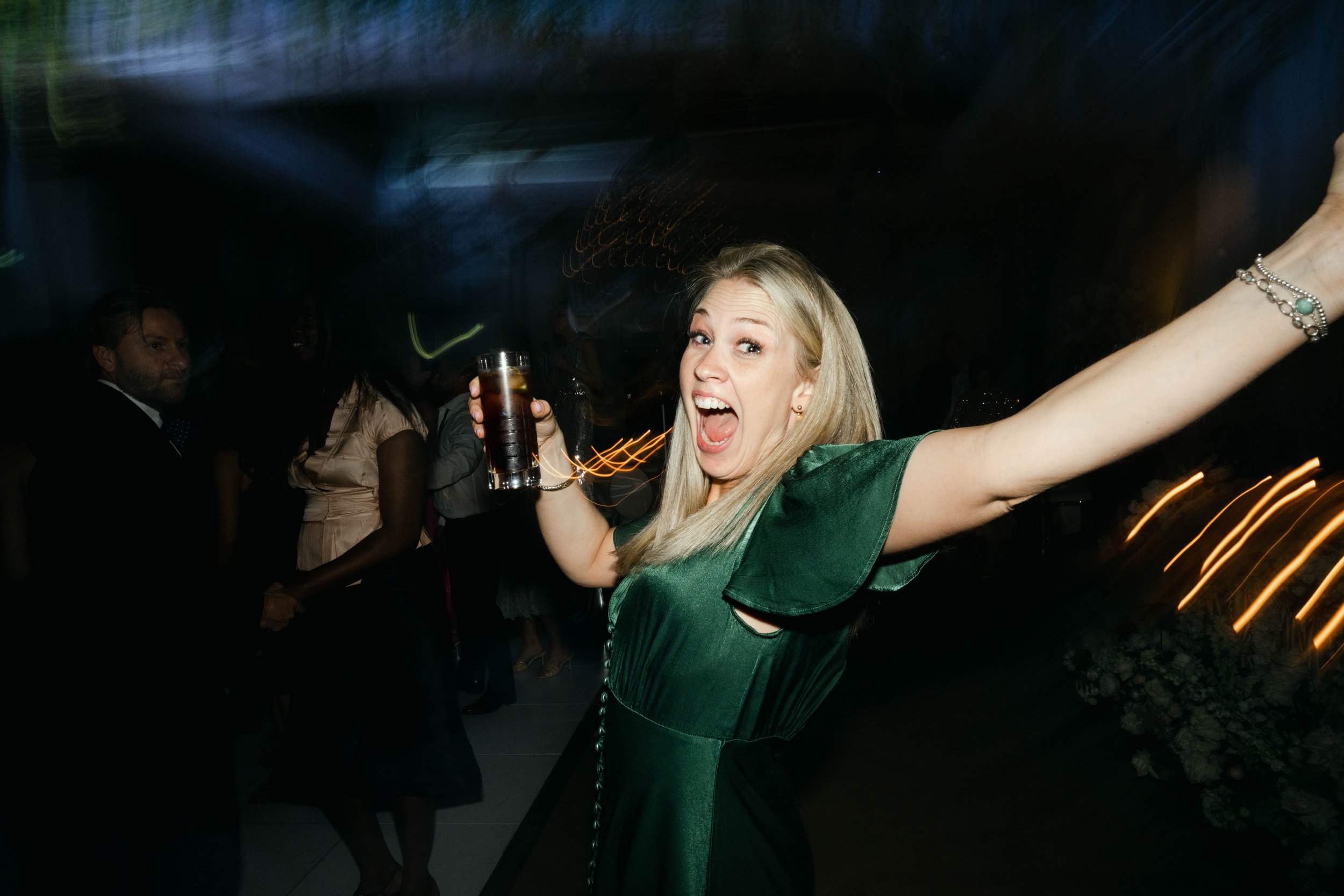 A woman in a green dress celebrating with a drink in a dimly lit party setting, smiling and raising her arm.