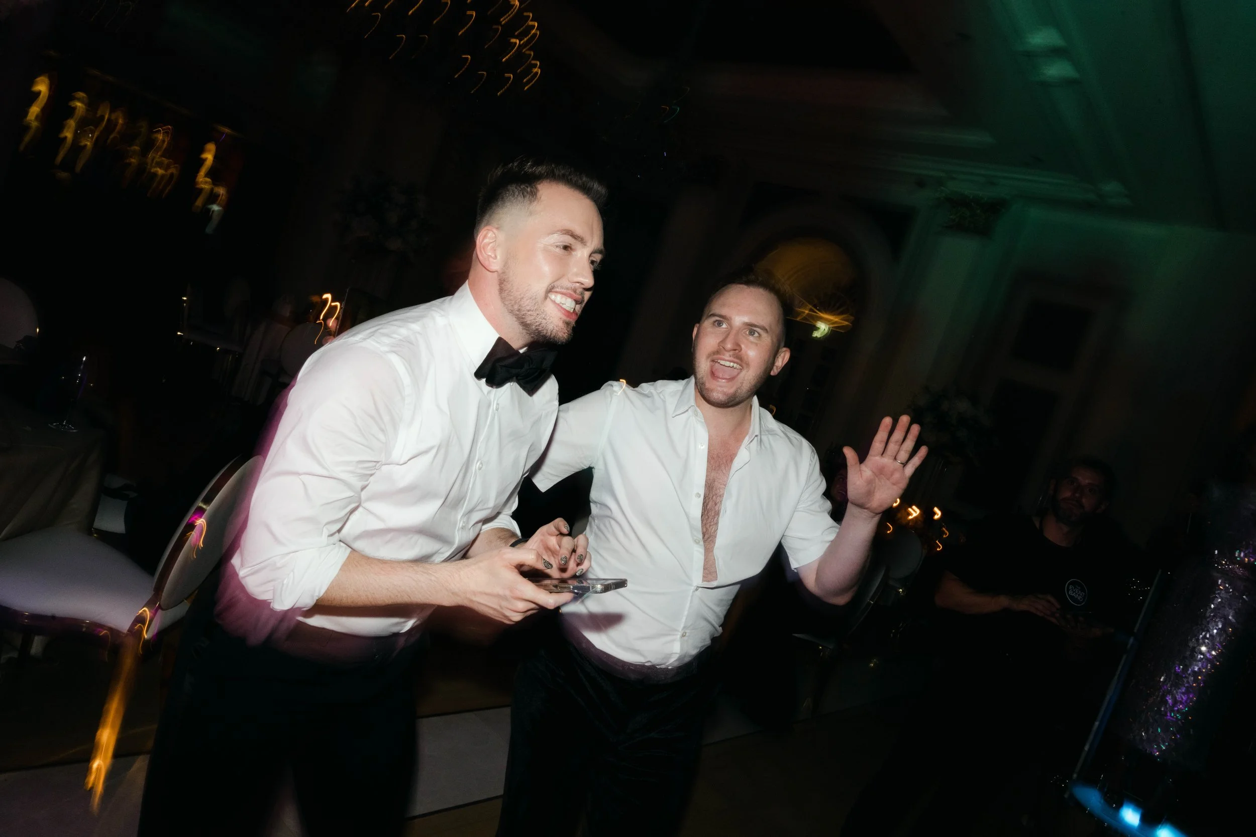 Two men at a party, one wearing a white shirt with rolled-up sleeves and a black bow tie, the other in a white unbuttoned shirt, are smiling and having fun. The man in the bow tie is holding a phone, and the other man is waving his hand. The backgrou