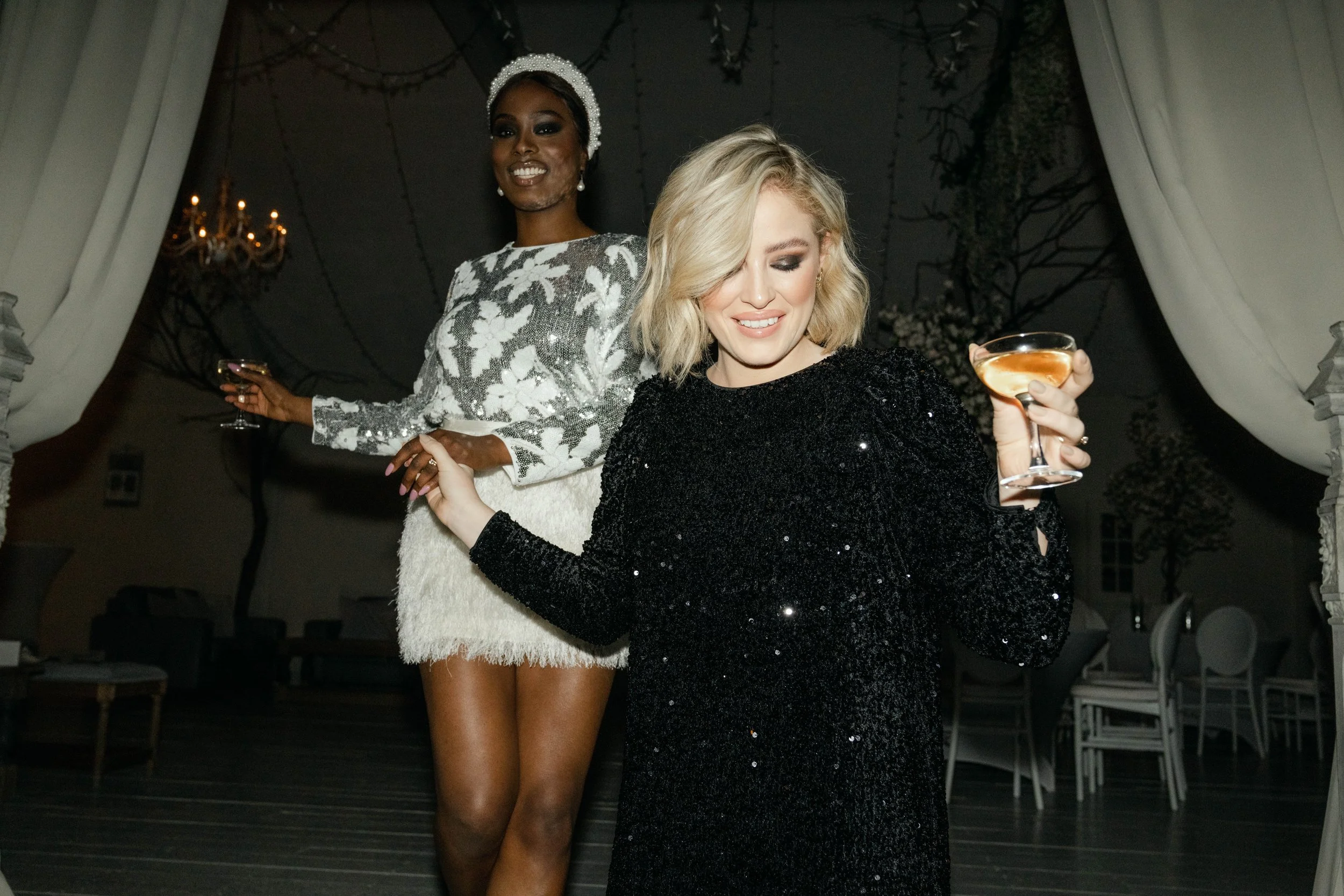 Two women in elegant attire holding drinks, smiling and dancing at a formal event.