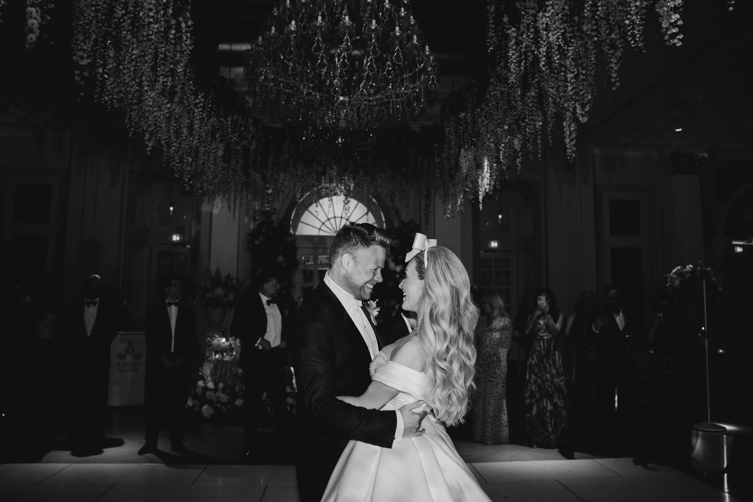 Black and white photo of a bride and groom sharing a dance at their wedding reception, surrounded by guests and elegant floral decorations.