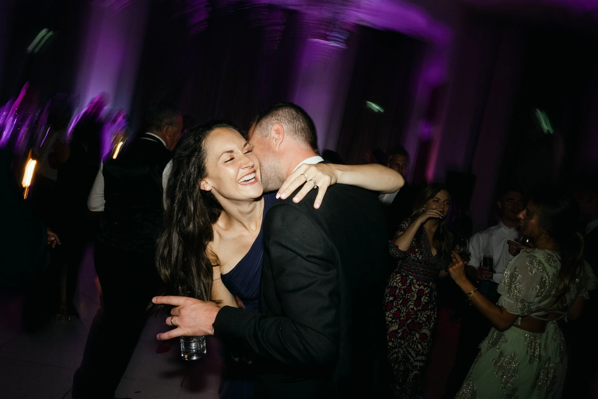 A joyful couple dancing and embracing at a party, surrounded by other guests, with purple lighting in the background.