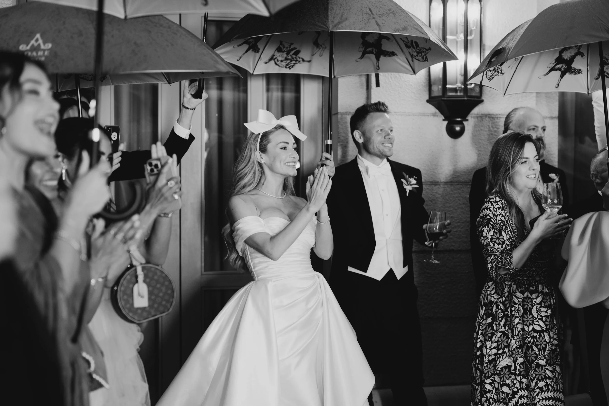 Black and white photo of a wedding reception with guests holding umbrellas, including a bride in a white wedding gown and a groom in a tuxedo, all smiling and enjoying the moment.
