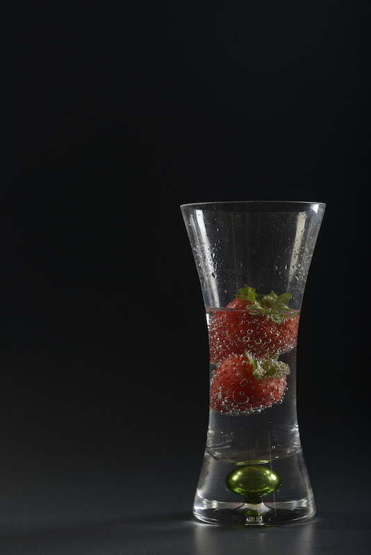 A glass vase filled with soda and strawberries sits in front of a solid black cardboard backdrop as part of a glass work project completed October 3, 2014. Soft boxes sit on either side of the object with a snoot above the vase to light the rim. 