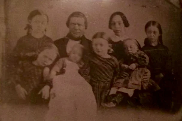 Norman and Jane Rundles with their children. This photo was taken shortly before they left New York by covered wagon for Iowa in 1856. Norman placed his sister's baby on his lap for the picture, as he wanted a way of visually marking Jane's pregnancy. Jane was pregnant during their journey by covered wagon and had their first son, named Norman, after his father, soon after they settled on the farm. Norman Jr. passed away at almost 3 months of age, and one of the pines was planted in his memory. The other pines were planted for three of the girls, who passed away quite suddenly from an epidemic that swept through the area during the winter of 1865-66.