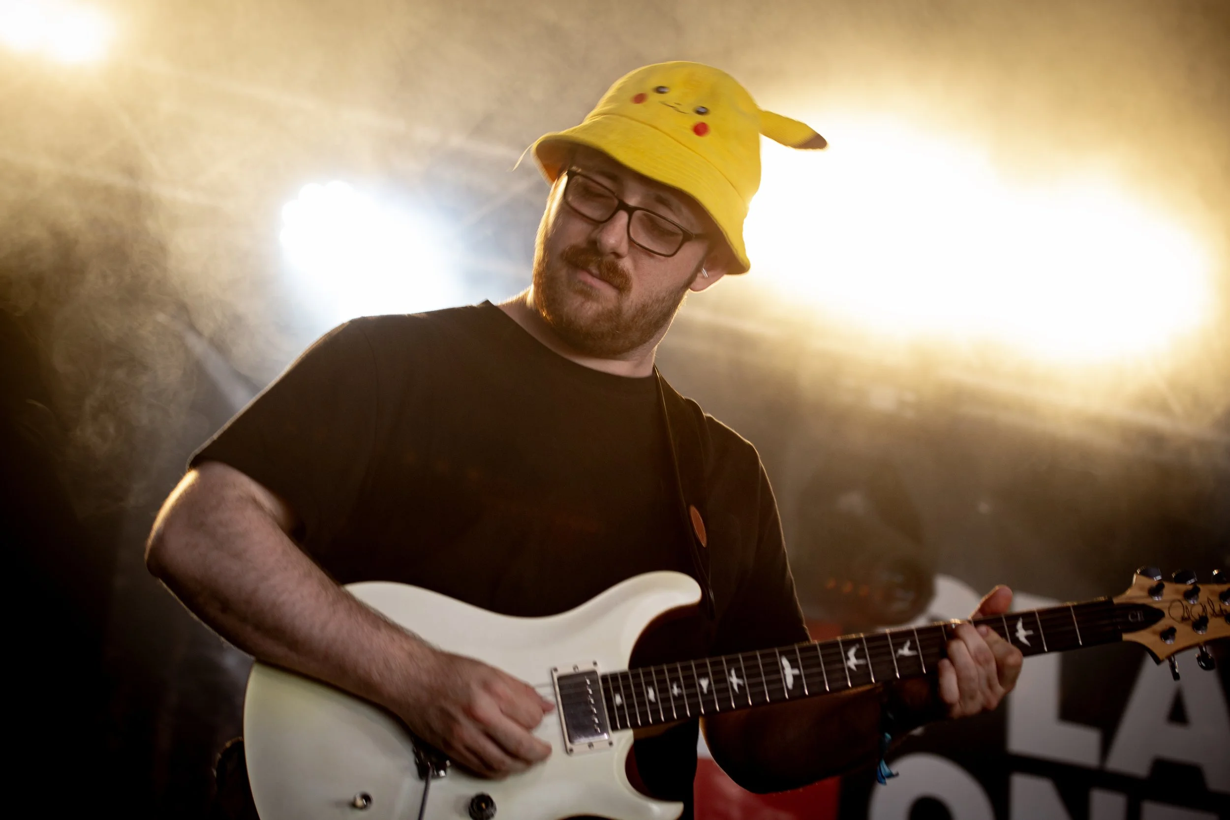 Joe Thompson playing guitar on stage  playing the Platform one stage at the Isle of Wight festival, wearing a yellow Pikachu hat, with stage lights in the background.