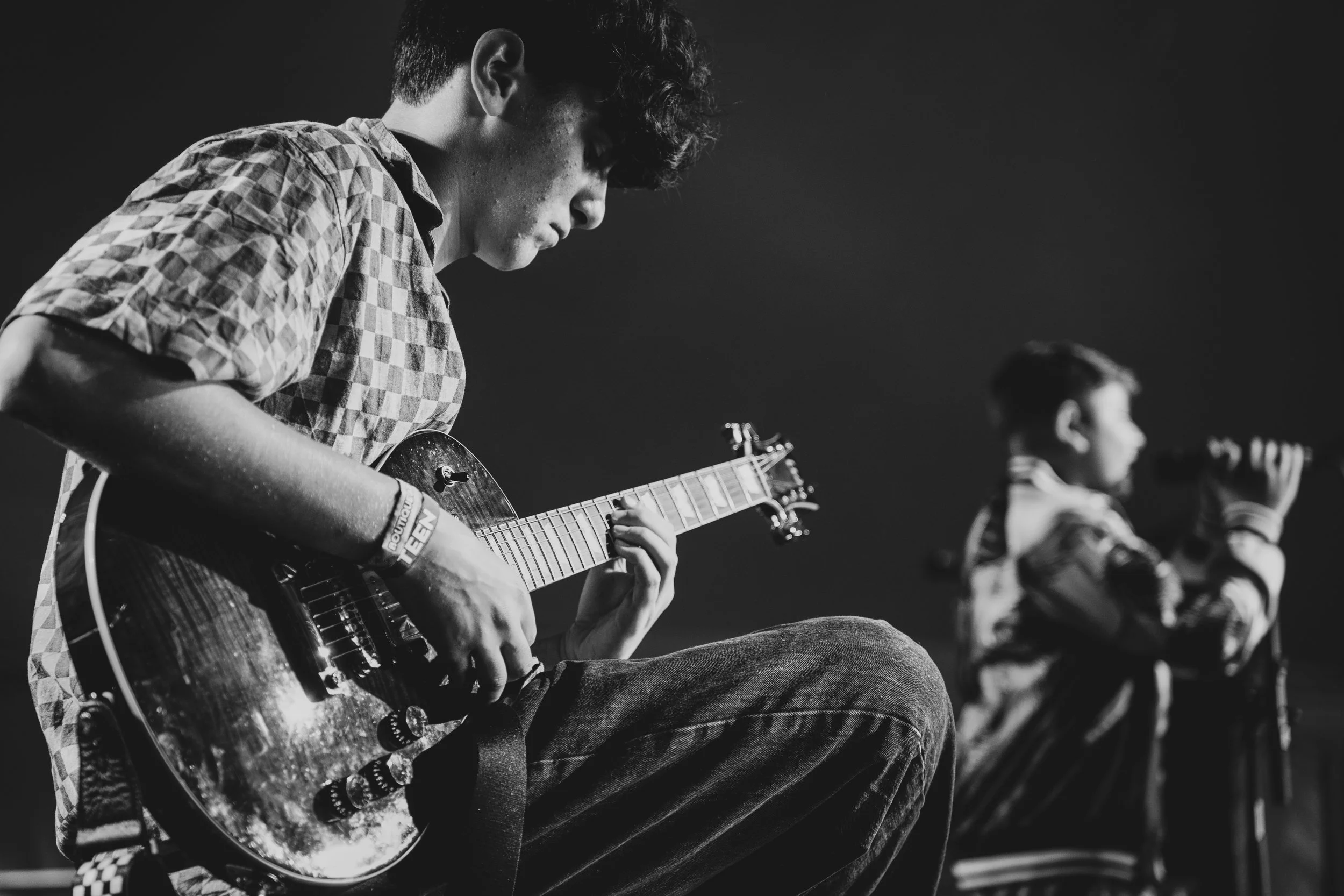 A young man playing an electric guitar on stage with another young man holding a microphone in the background.