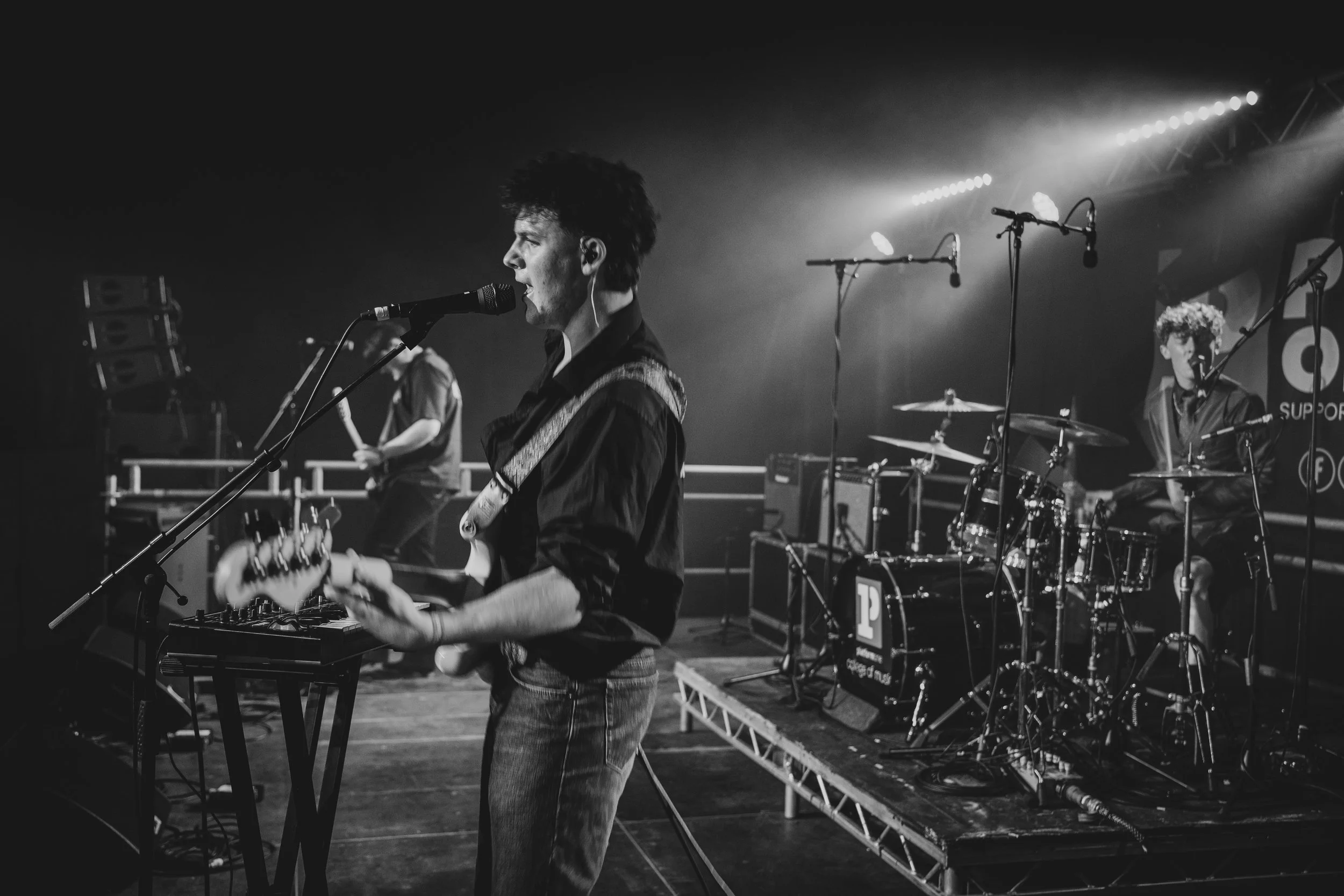 Black and white photo of a band performing on stage, with a guitarist and vocalist in the foreground, and a drummer and second vocalist in the background, all surrounded by musical equipment.