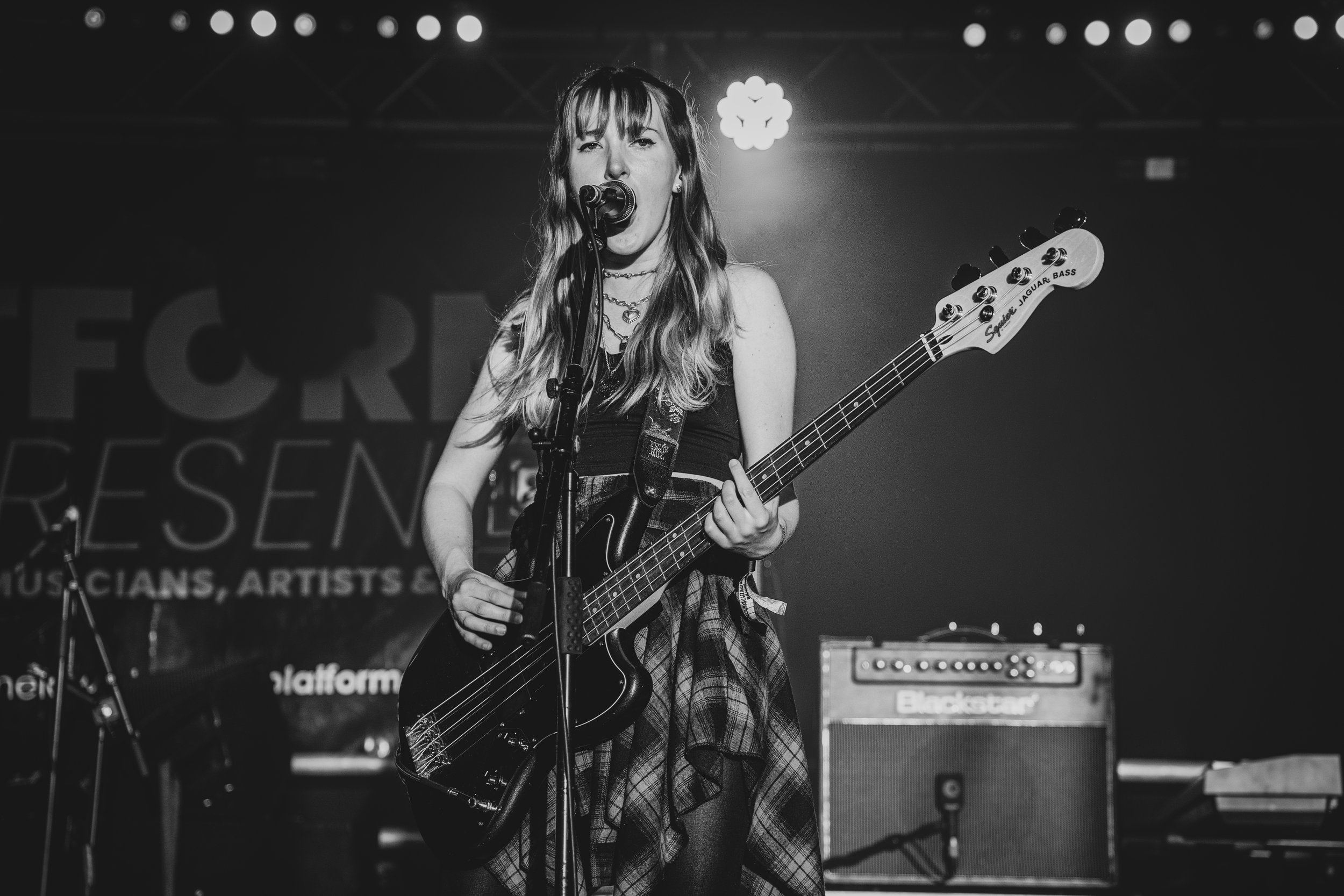 A female musician singing into a microphone while playing an electric bass guitar on stage in black and white.