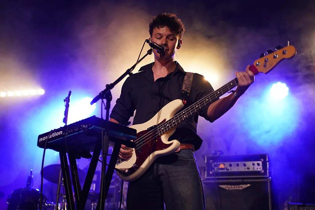 Young man performing on stage with a bass guitar, singing into a microphone, with colorful stage lighting and musical equipment in the background.