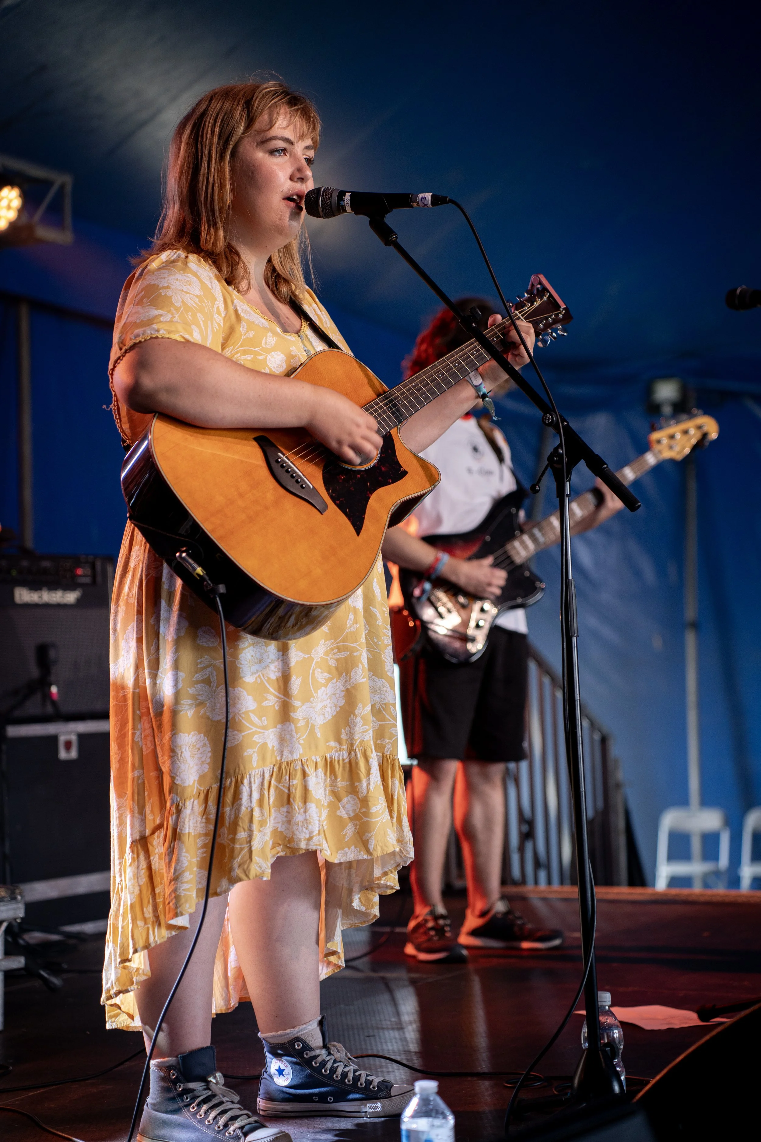 Amy Joliffe playing the Platform one stage at the Isle of Wight festival, with red hair in a yellow floral dress playing an acoustic guitar and singing into a microphone on stage, with a bassist in the background.