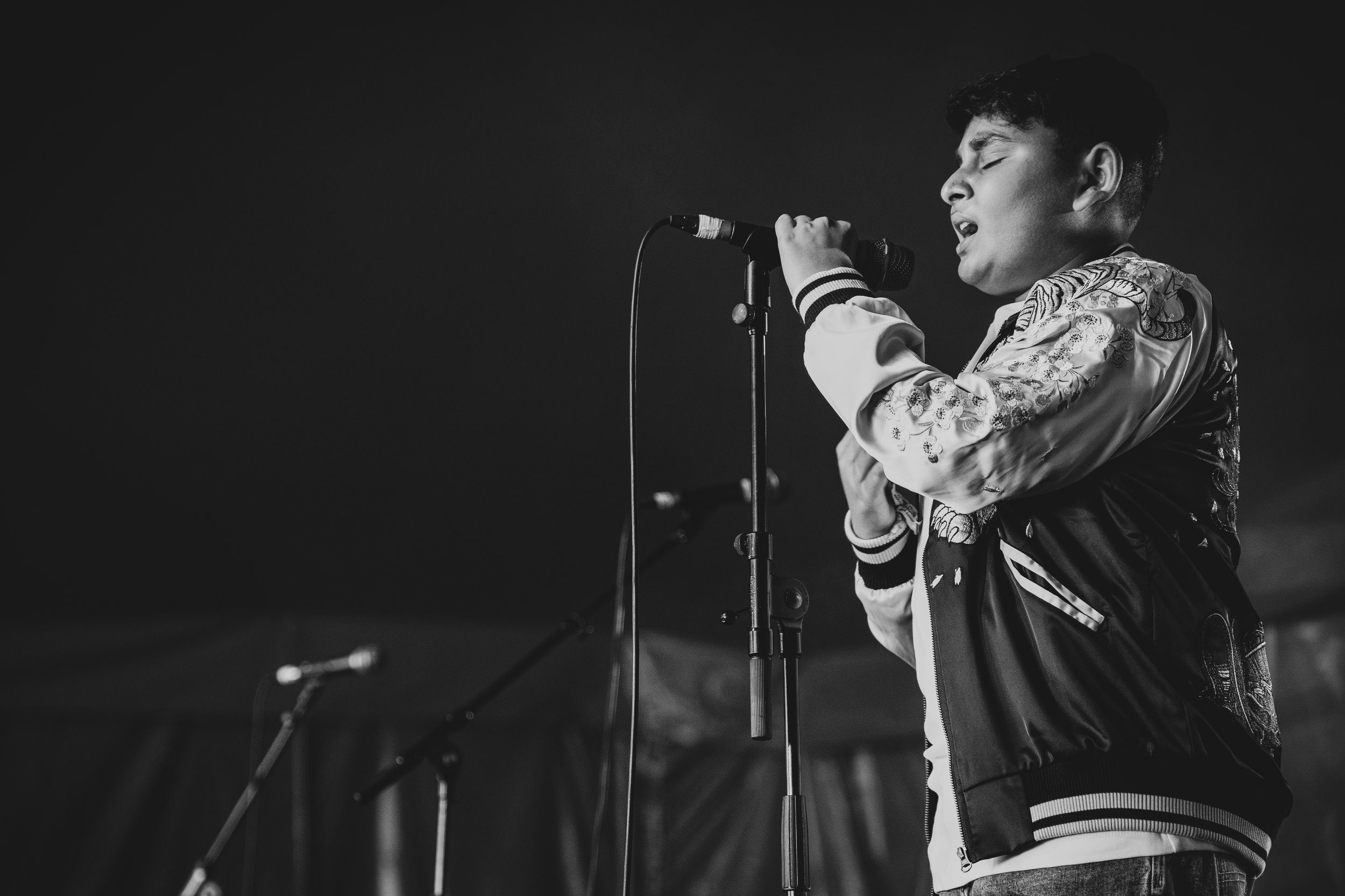 A young man singing into a microphone on stage, with closed eyes and wearing a jacket with floral embroidery.
