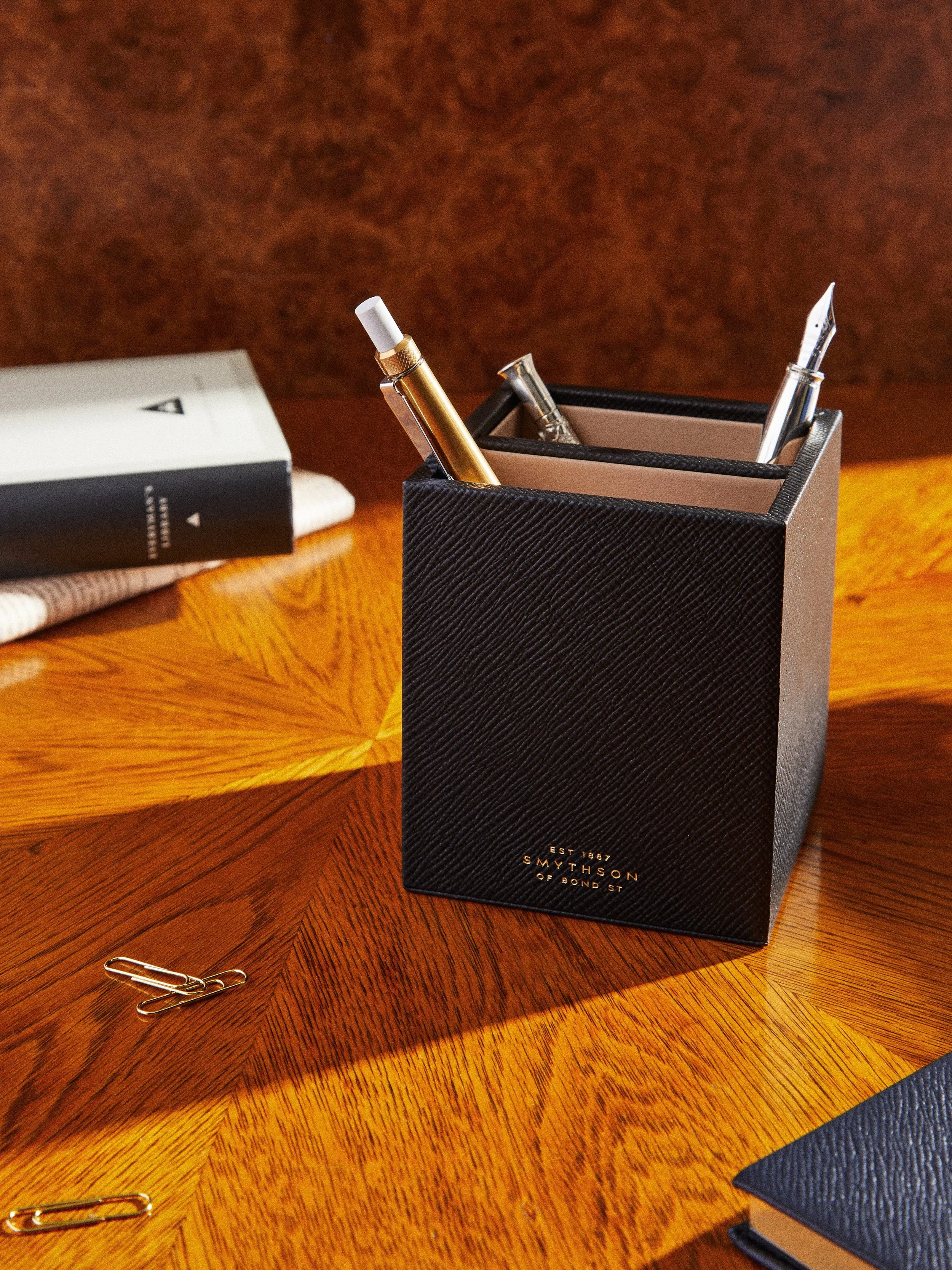 A black textured organization holder containing pens on a wooden desk, with a gold paperclip and a folded paper in the background.