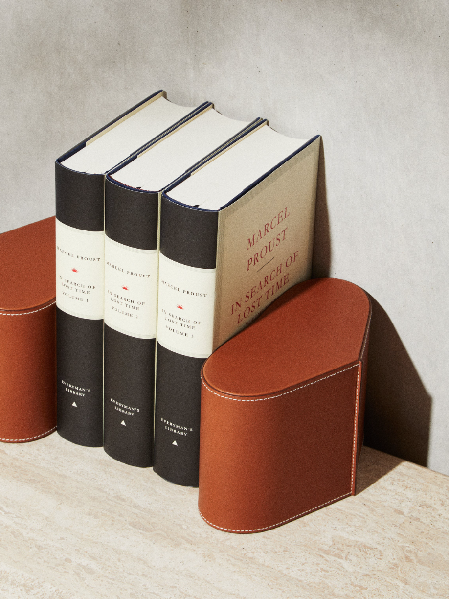 Three books titled 'In Search of Lost Time' by Marcel Proust are standing upright next to a brown leather bookend shaped like a semicylinder with a curved top. The books have black and white covers with red detailing, and the bookend has white stitch