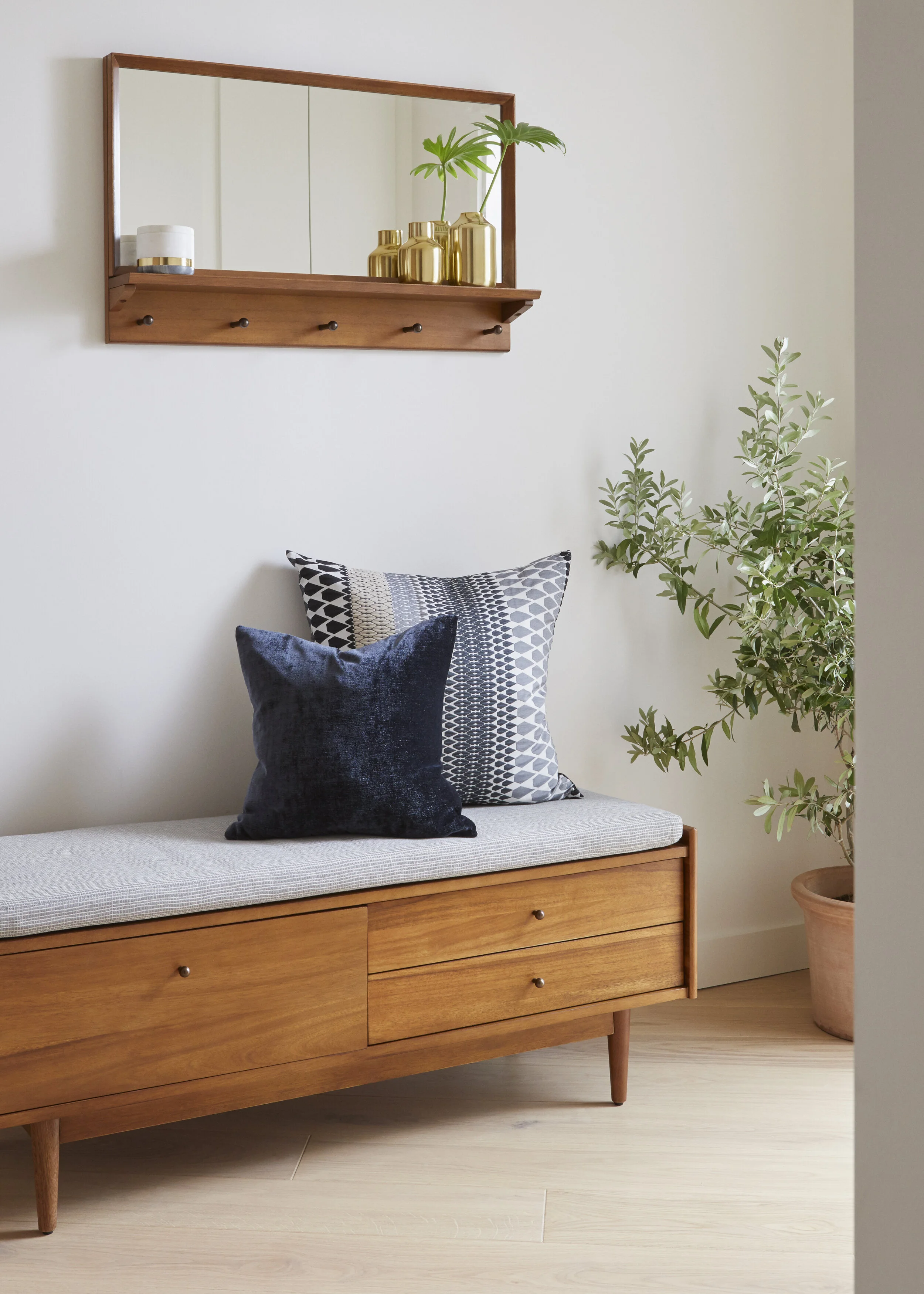 Minimalist entryway or hallway corner with a wooden bench, two decorative pillows, a potted plant, and a wall-mounted mirror with a wooden frame above. The mirror has a shelf with gold decorative vases and small candles.