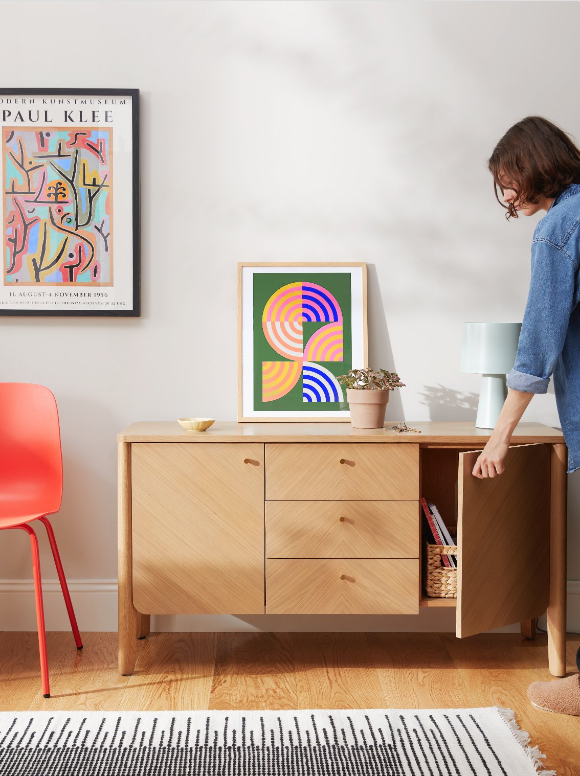 A woman opening a wooden cabinet in a living room or gallery space with framed artwork on the wall and decorative items on top of the cabinet.