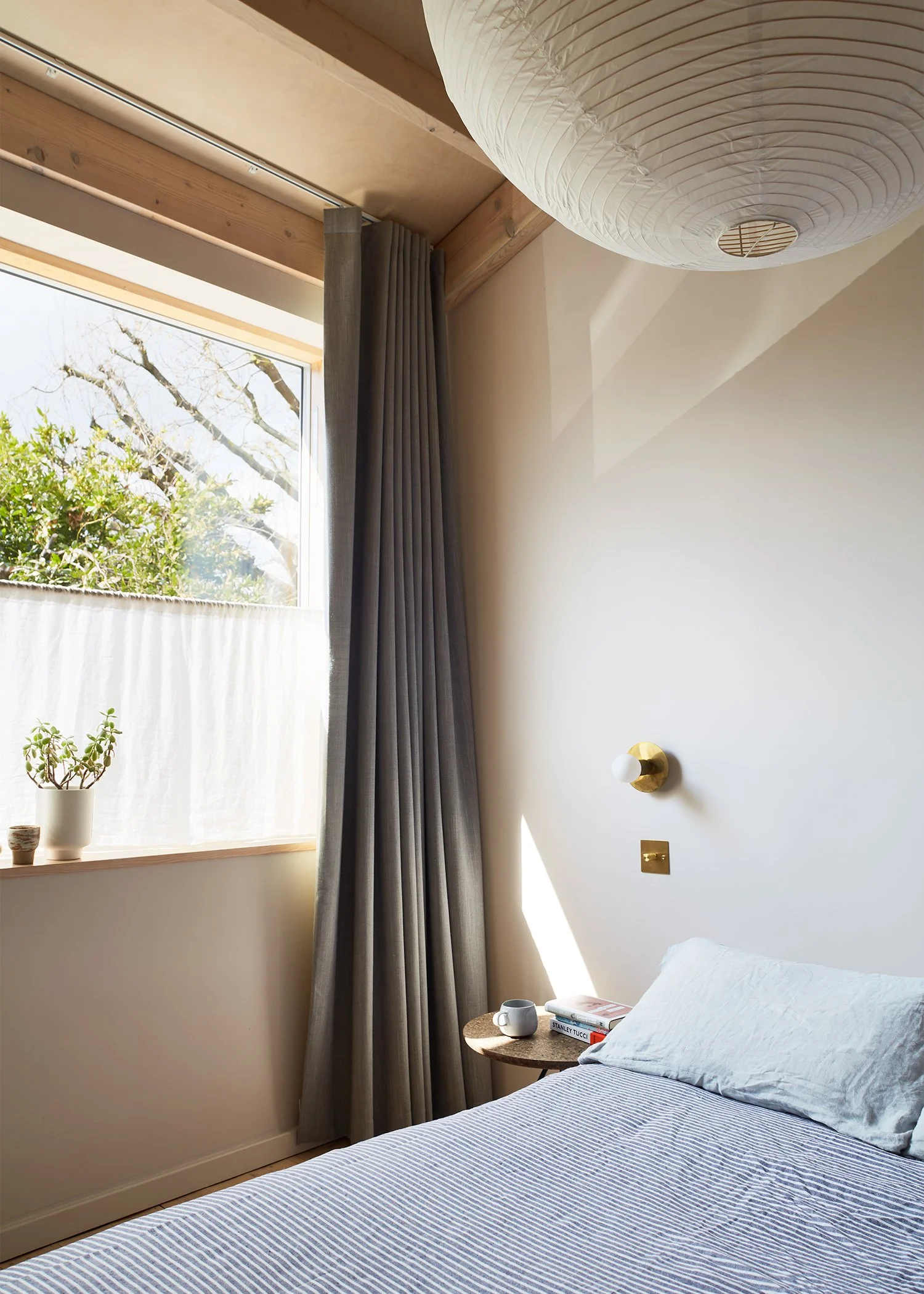 A cozy bedroom with a bed, a small side table with a coffee cup and books, a window with a plant on the sill, and a paper lantern ceiling light.
