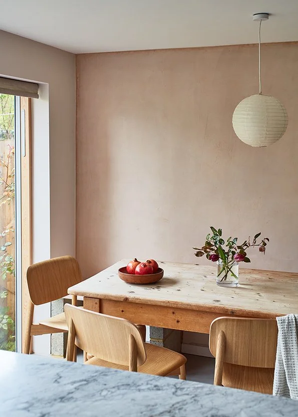 A dining area with a wooden table, four light wooden chairs, a bowl of apples, and a glass vase with flowers. A paper lantern hangs from the ceiling, and there is a window with a roller shade letting in natural light.