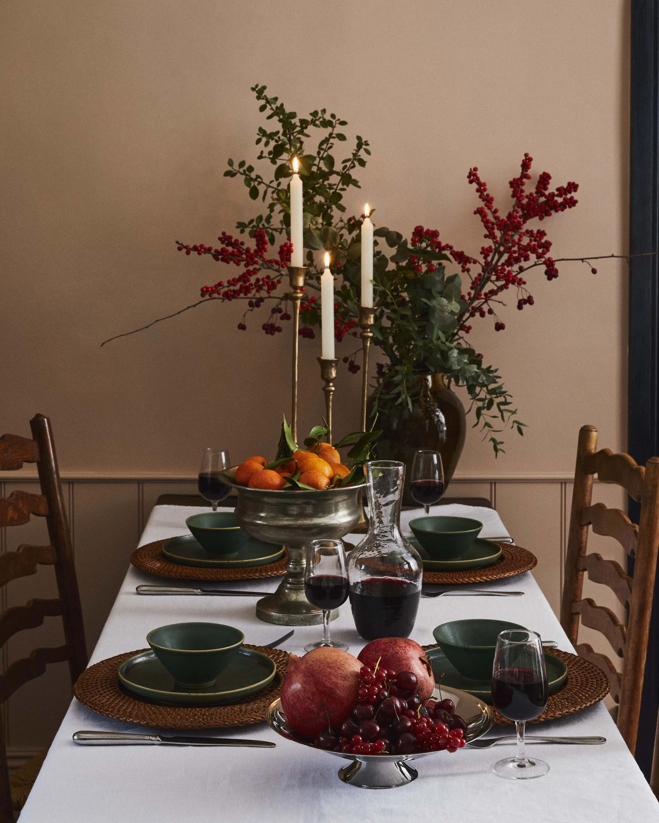 A festive dining table with a white tablecloth, green dishes, and glassware containing red wine. A large silver bowl and a silver platter hold pomegranates, grapes, and red berries. Tall candlesticks with lit white candles and a large vase with green