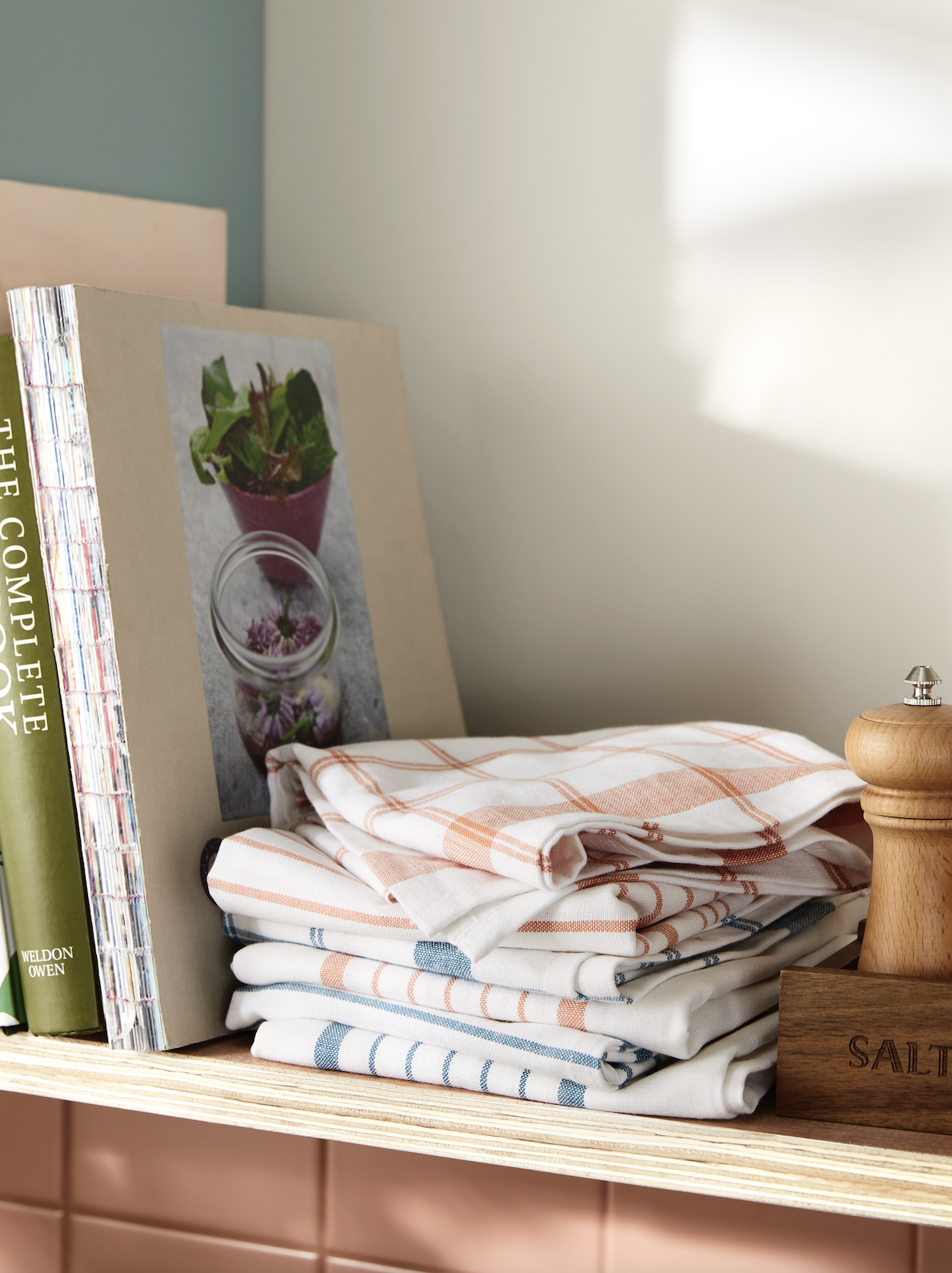 Stacked dish towels with orange and blue stripes on a kitchen shelf, a cookbook leaning against the wall, and a wooden salt grinder nearby.
