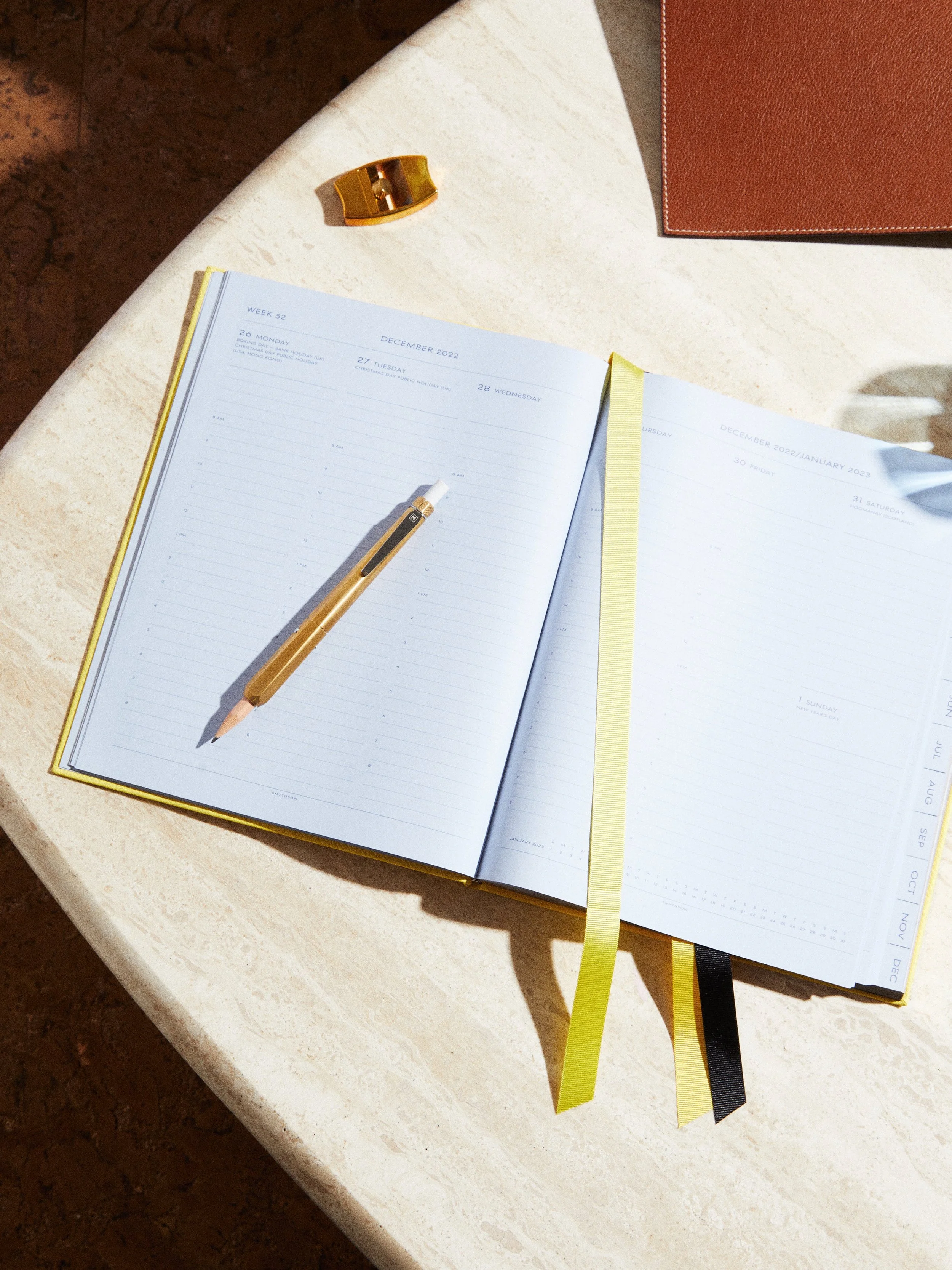Open planner for December 2022 on a white marble table with a yellow and a black bookmark, a gold pen, a gold paper clip, and a brown leather item.