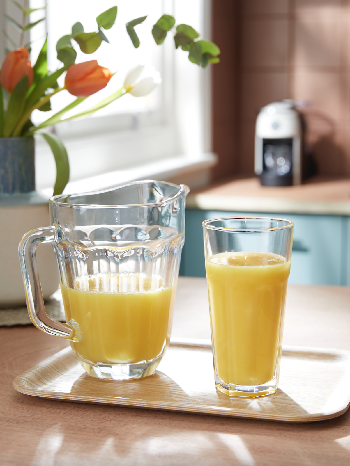 Glass pitcher and glass filled with orange juice on a tray on a kitchen table, with flowers and a coffee maker in the background.