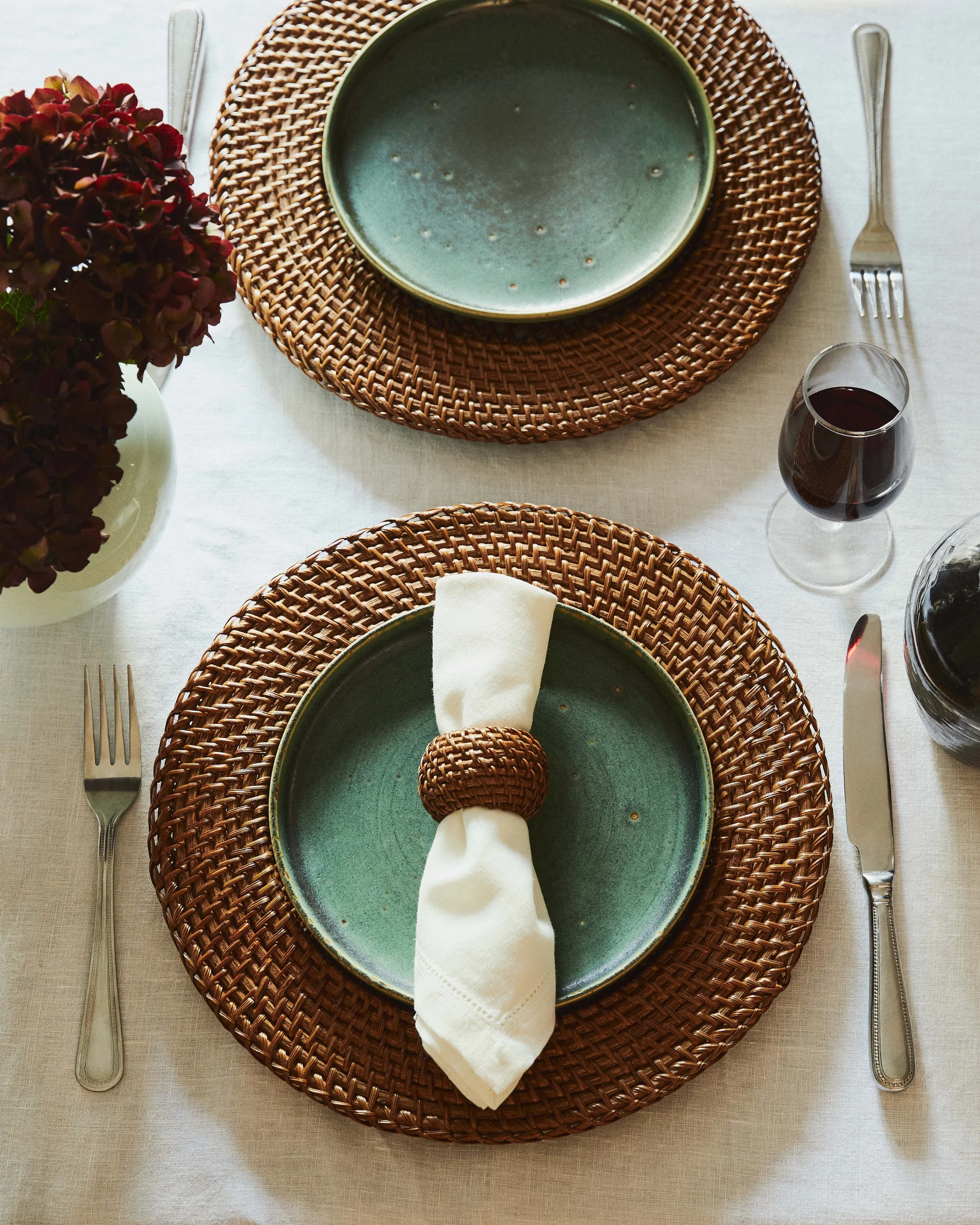 Place setting with green ceramic plate on a woven charger, napkin held with a woven ring, and cutlery on a white tablecloth, along with a glass of red wine and a vase of burgundy flowers.