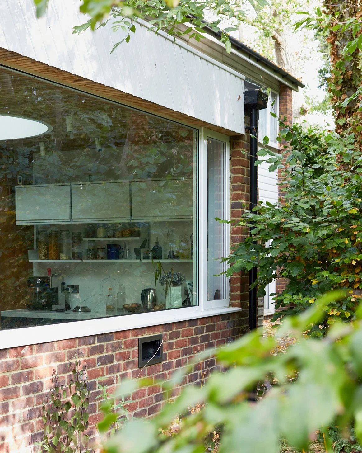Part of a brick house with a large window showing a kitchen interior, surrounded by green foliage.
