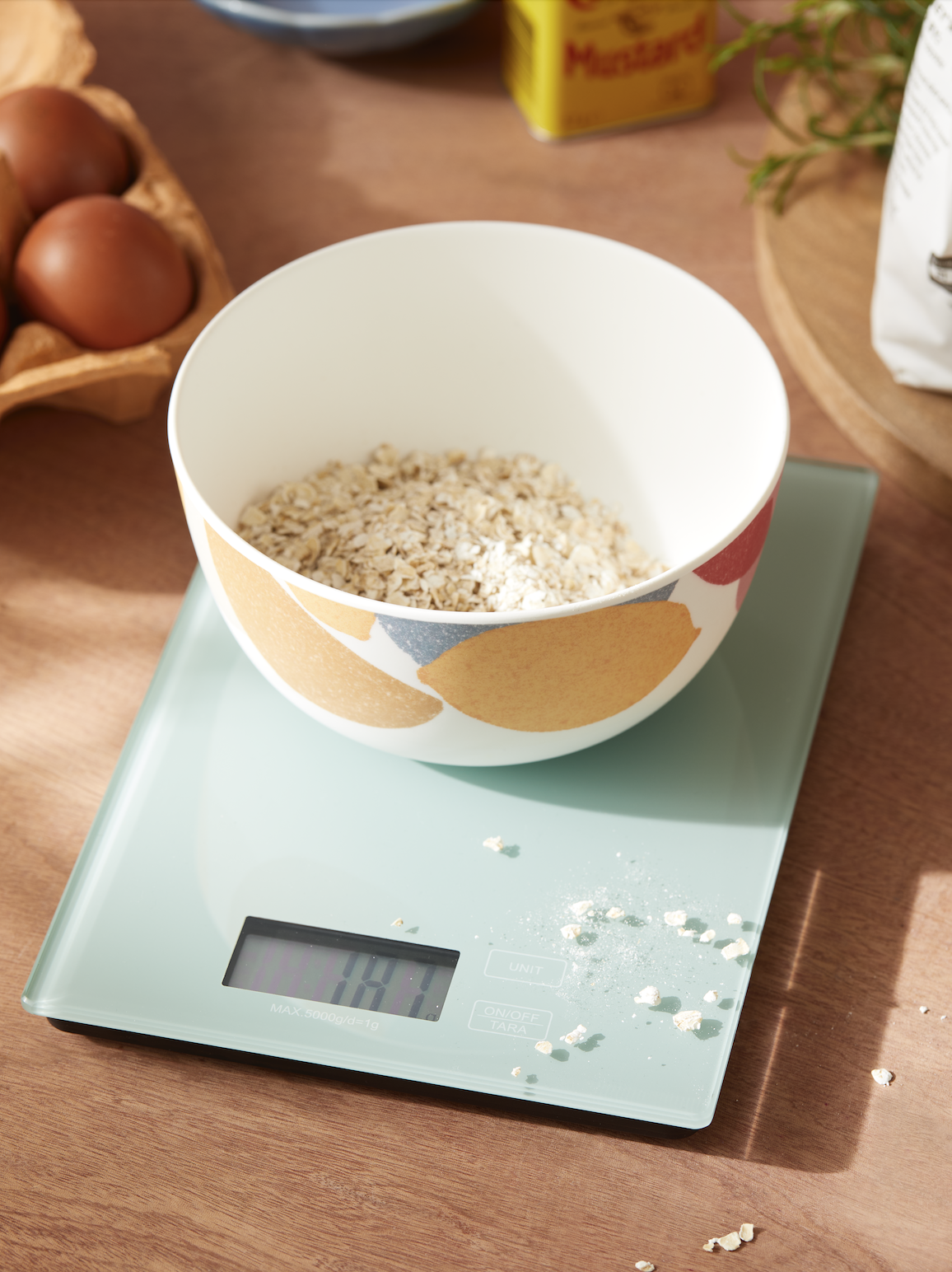 A bowl with a colorful fruit pattern on a digital kitchen scale with some oats spilled around, on a wooden surface.