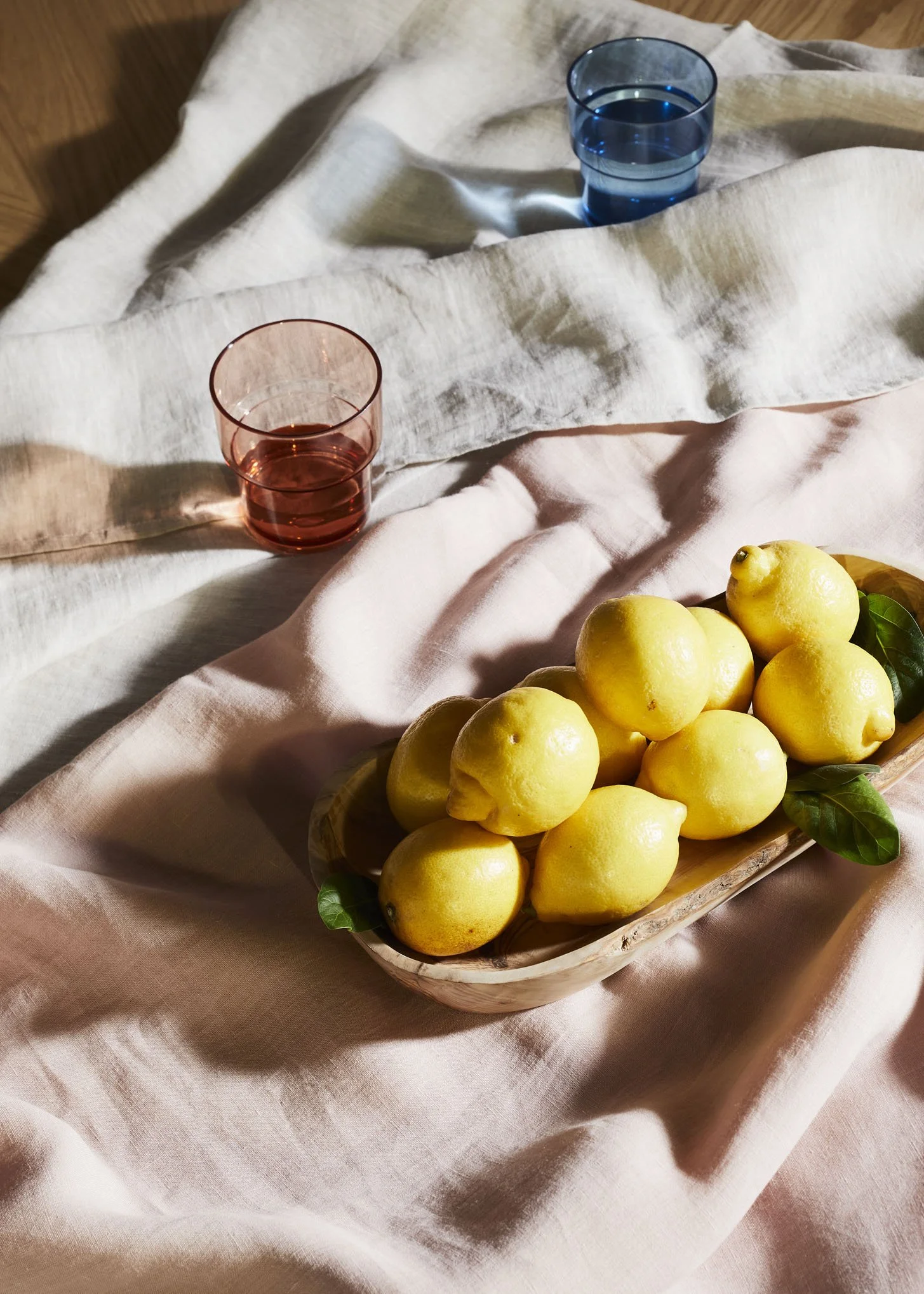 A wooden bowl filled with yellow lemons on a pink cloth, with two glasses, one blue and one pink, on a layered fabric surface.