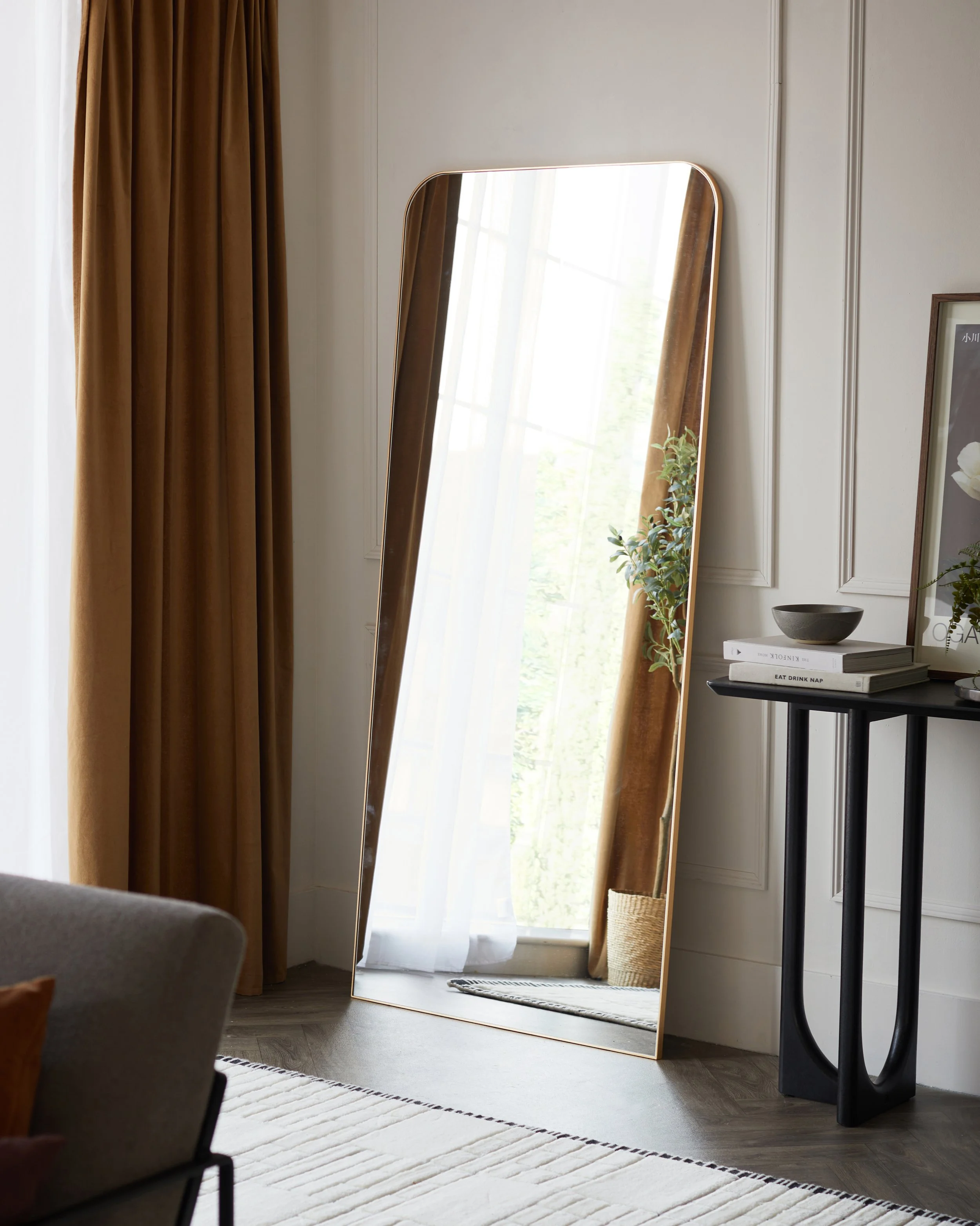 A tall, leaning mirror with a gold edge reflects a window with sheer white curtains and brown drapes, a potted plant, and a sunlight-lit room.