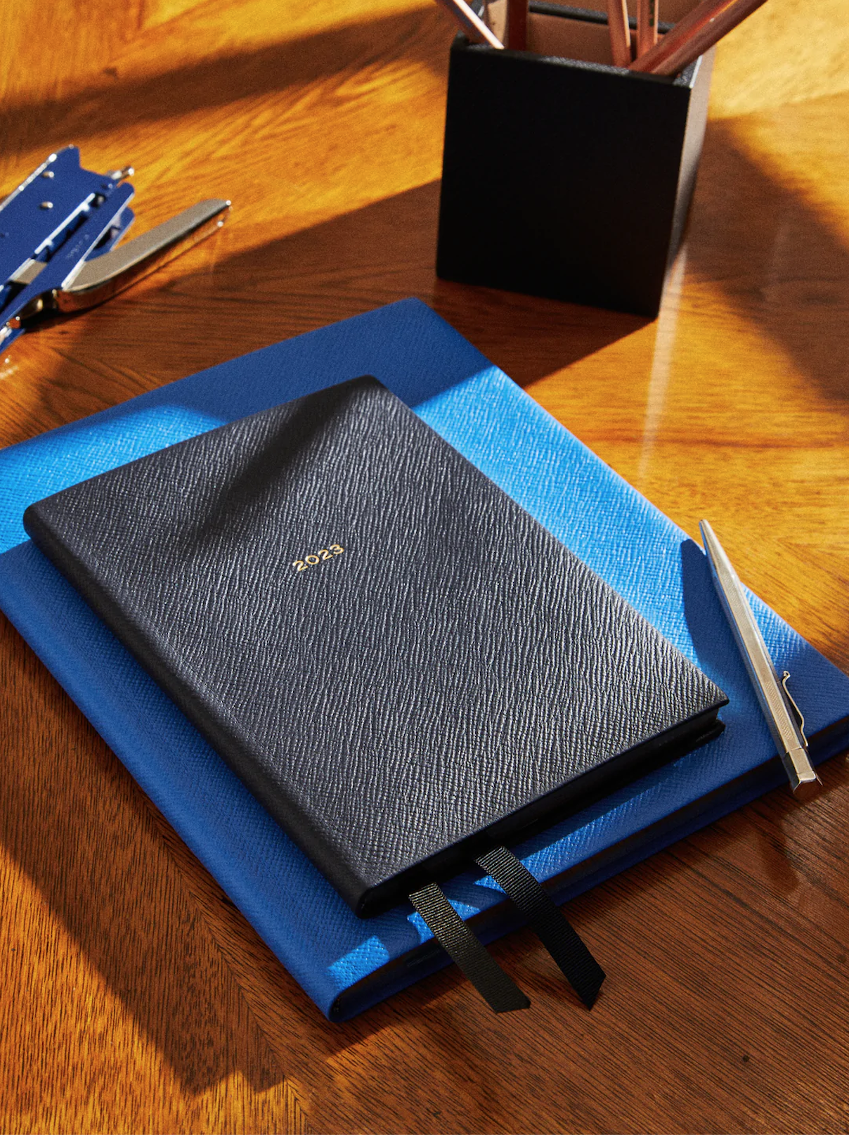 Black 2023 planner on a blue notebook, a silver pen, a black pen holder, and a stapler on a wooden desk.