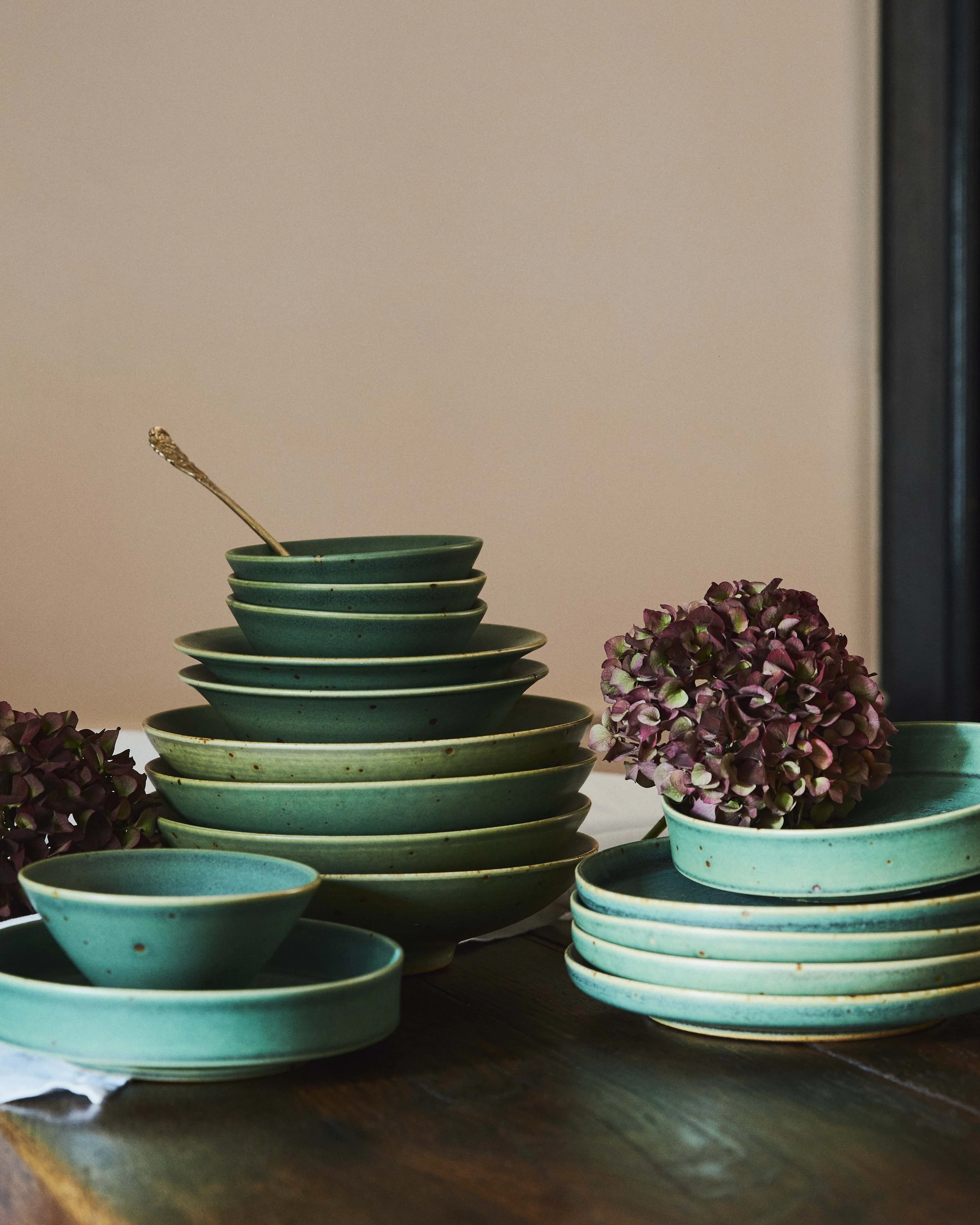 Stacked green ceramic bowls and plates on a wooden table, decorated with purple hydrangea flowers.