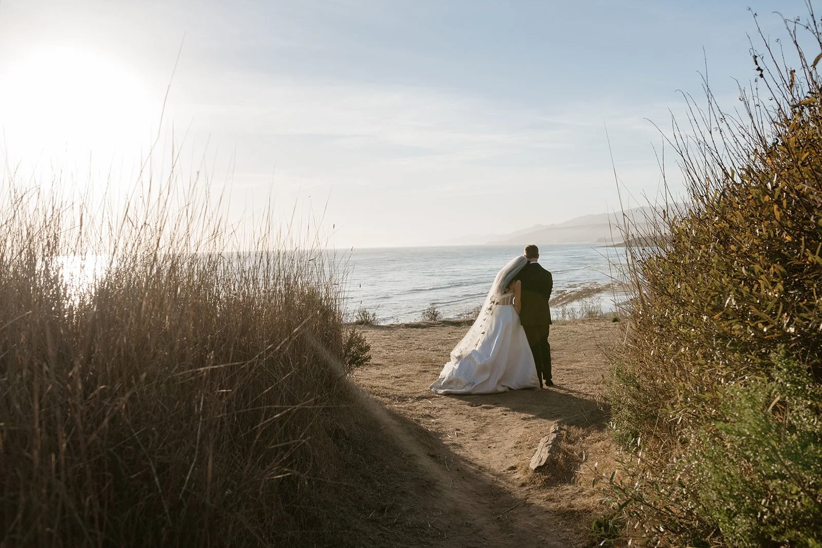 A bride and groom walking on a dirt path towards the ocean, surrounded by dry bushes and grass, with a sunset sky in the background.