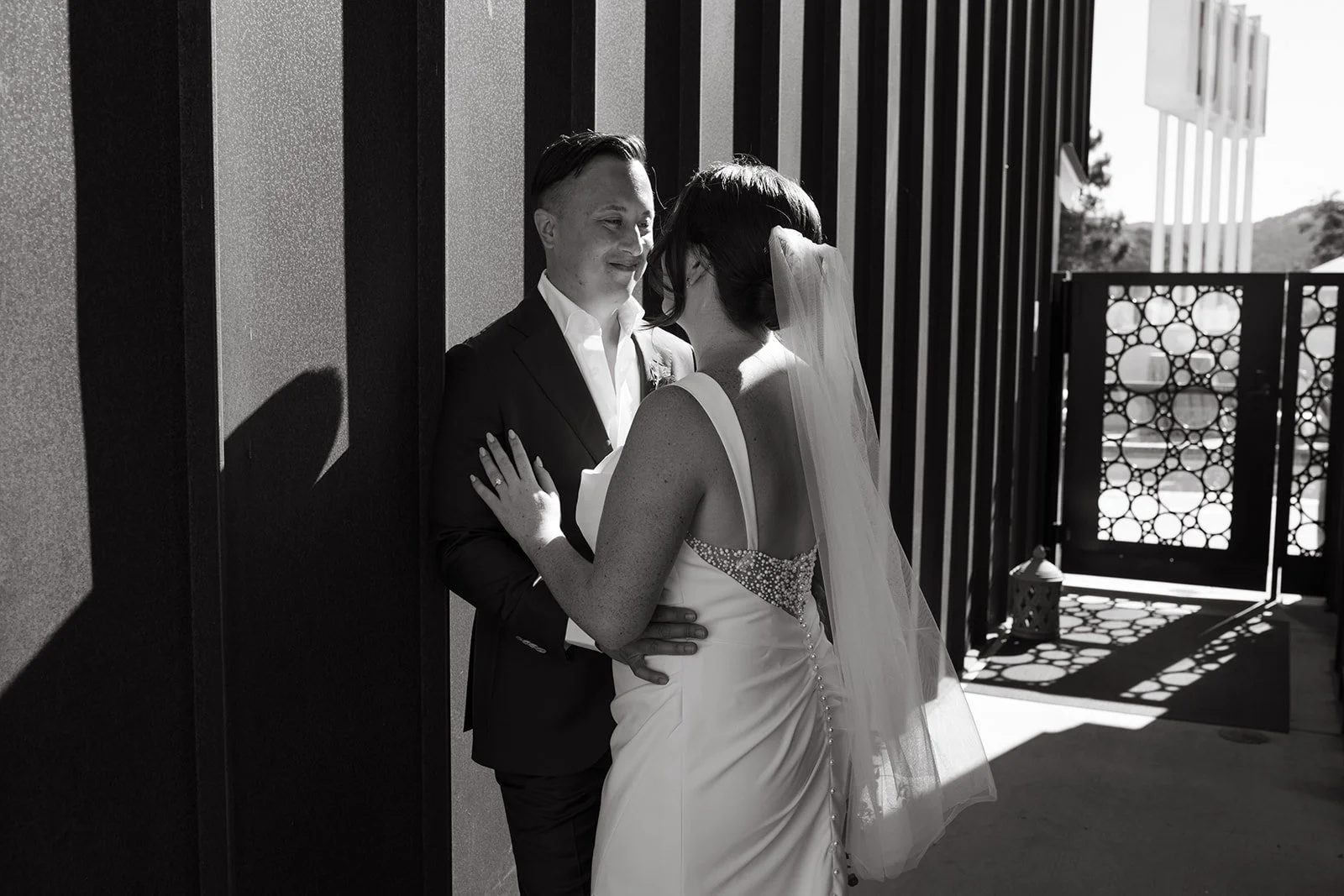 A black and white photo of a bride and groom on their wedding day, standing close to each other outdoors, smiling, with the bride wearing a wedding dress with a veil and the groom in a suit, positioned against a patterned fence with bright sunlight casting shadows.
