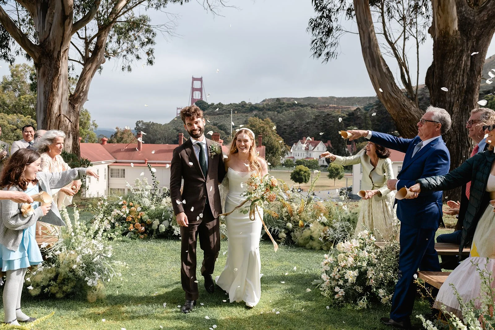 A wedding celebration outdoors with a bride and groom walking under a shower of flower petals, surrounded by family and friends, with the Golden Gate Bridge visible in the background.