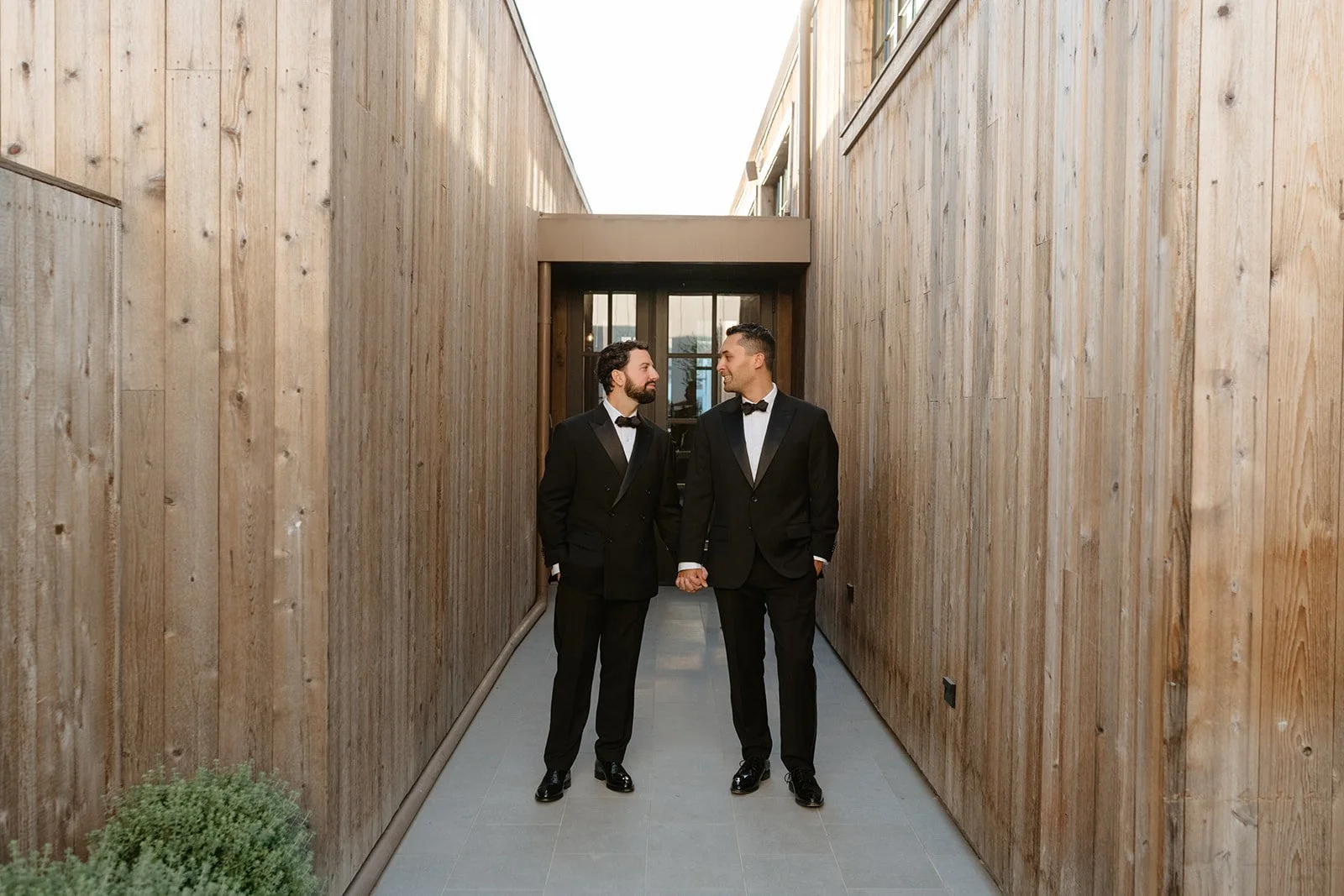 Two men dressed in black tuxedos holding hands and looking at each other in a narrow outdoor passage with wooden walls.