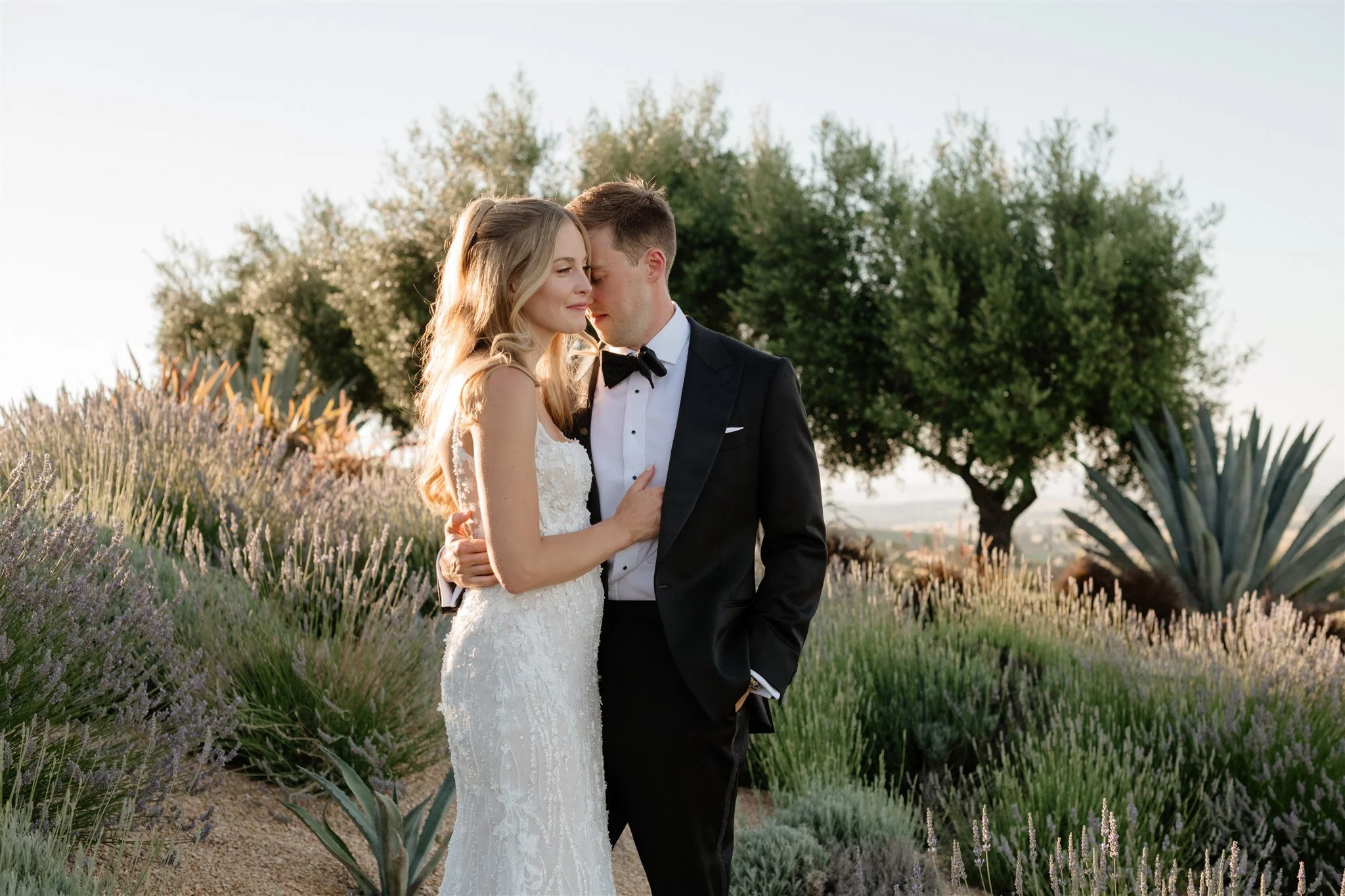A bride and groom stand closely together in a garden during sunset, with trees and plants in the background.