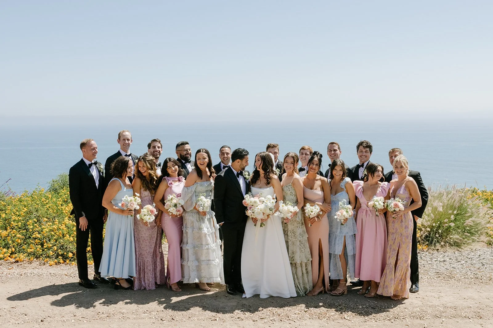 A large group of people dressed in wedding attire standing outdoors near the ocean, smiling for a photo on a sunny day.