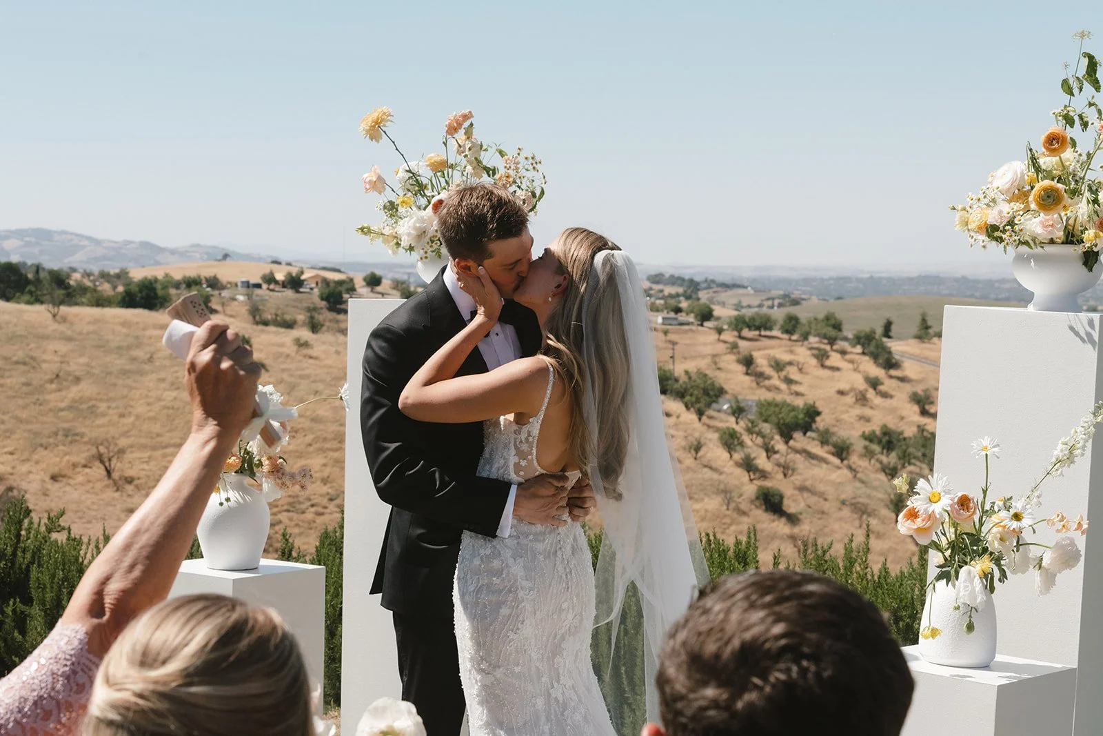 A bride and groom share a kiss at their outdoor wedding ceremony with scenic hills in the background, decorated with white and pastel flowers.
