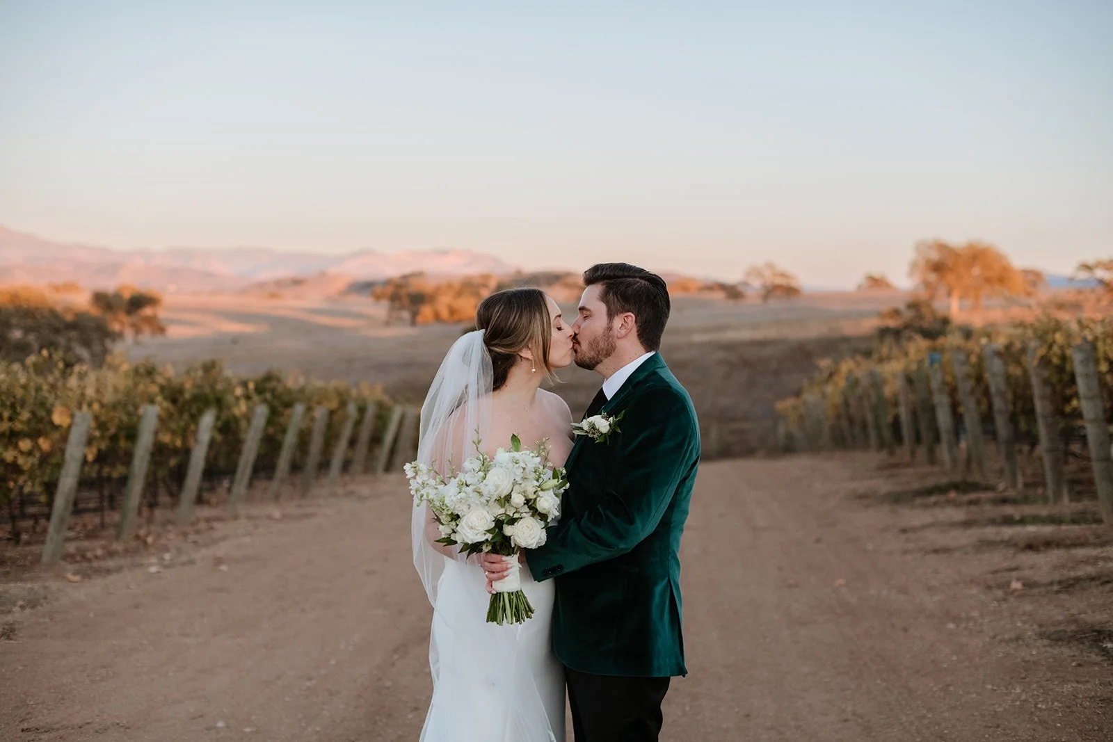 A bride and groom kiss in a vineyard at sunset, the bride holding a bouquet of white flowers.